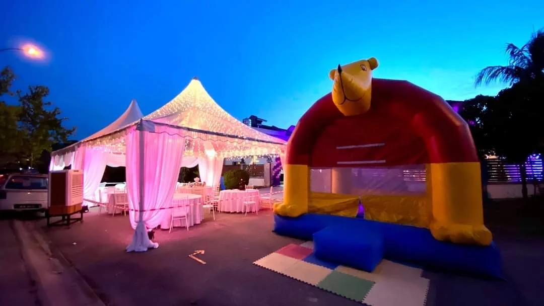 Pink illuminated outdoor event tent with tables and chairs, and a colorful inflatable bounce house shaped like a horse, set against a twilight sky.