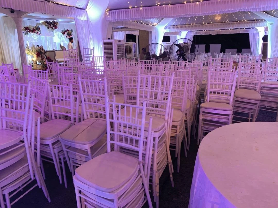 Indoor event space decorated with white chairs, floral arrangements, and purple lighting, with a stage area at the front and ceiling draped with fabric and string lights.