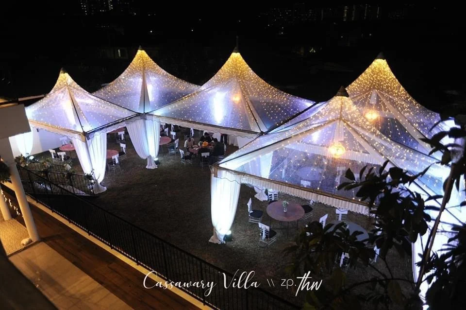 Outdoor event tent decorated with string lights and white drapes, set up for an evening gathering or celebration, seen from an elevated angle.