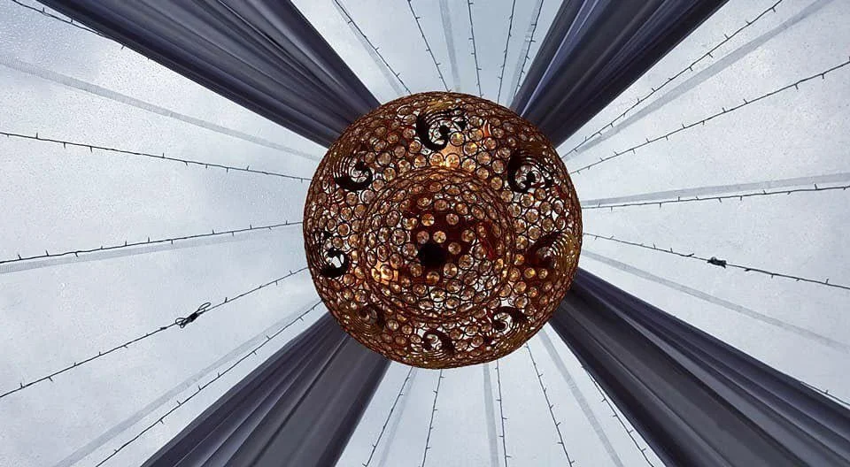 A spherical chandelier made of multiple circular metal rings hanging from a ceiling with fabric drapes and string lights.