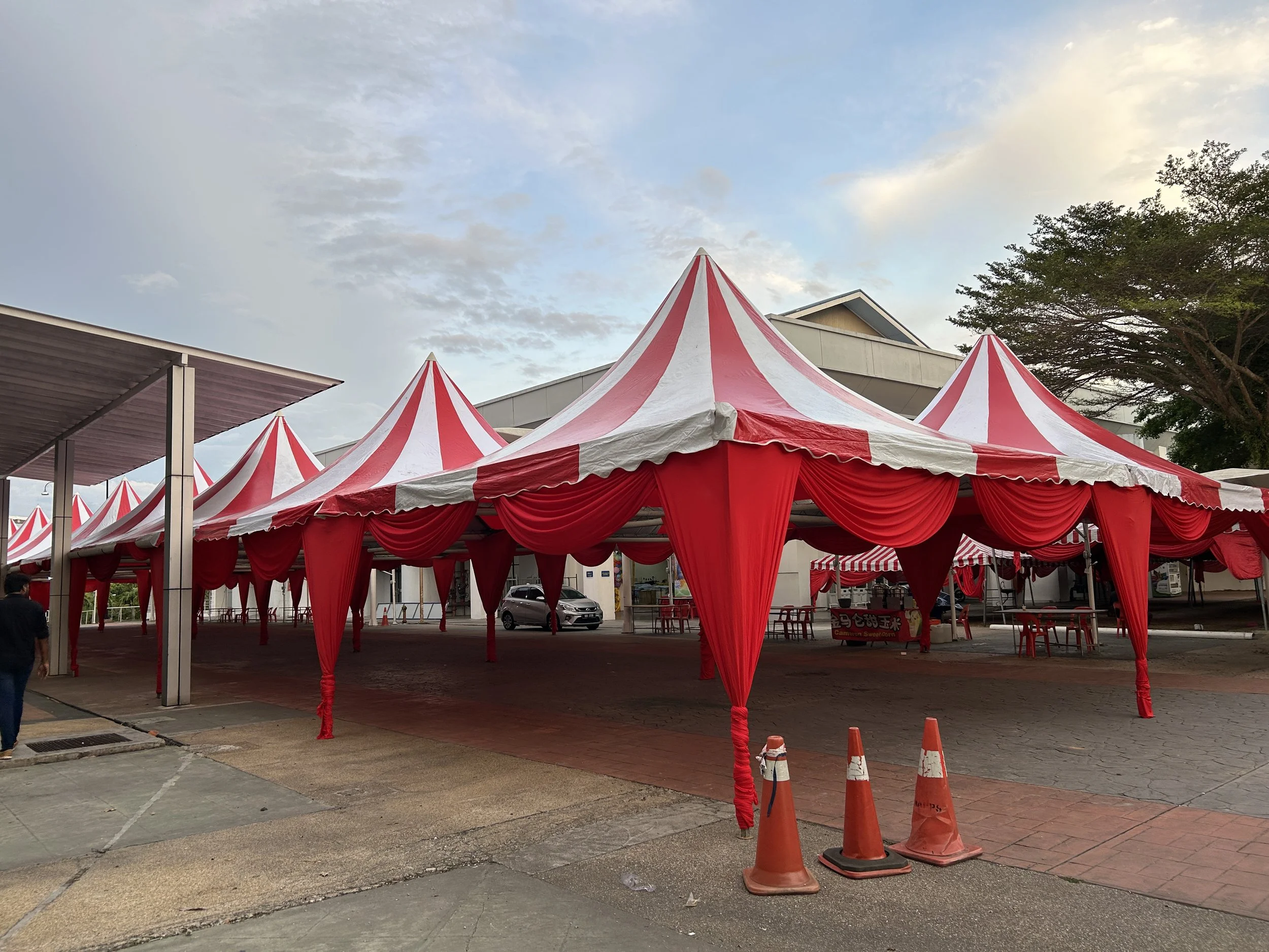 Red and white striped circus tents set up outdoors under a partly cloudy sky, with some trees and a building in the background.