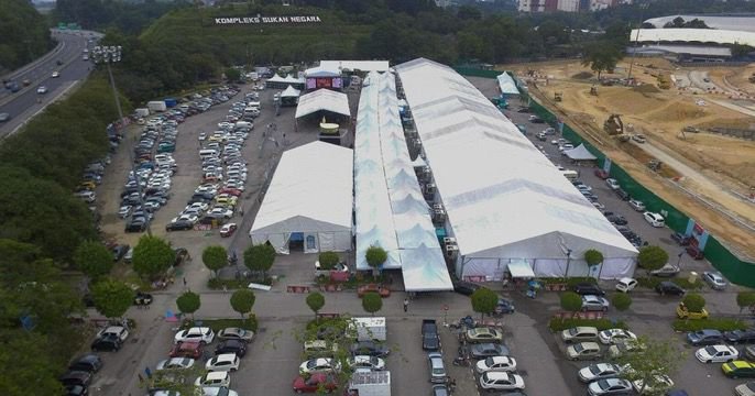 Aerial view of a large outdoor marketplace with multiple white tents, surrounded by parking lots filled with cars and some trees lining the walkways.