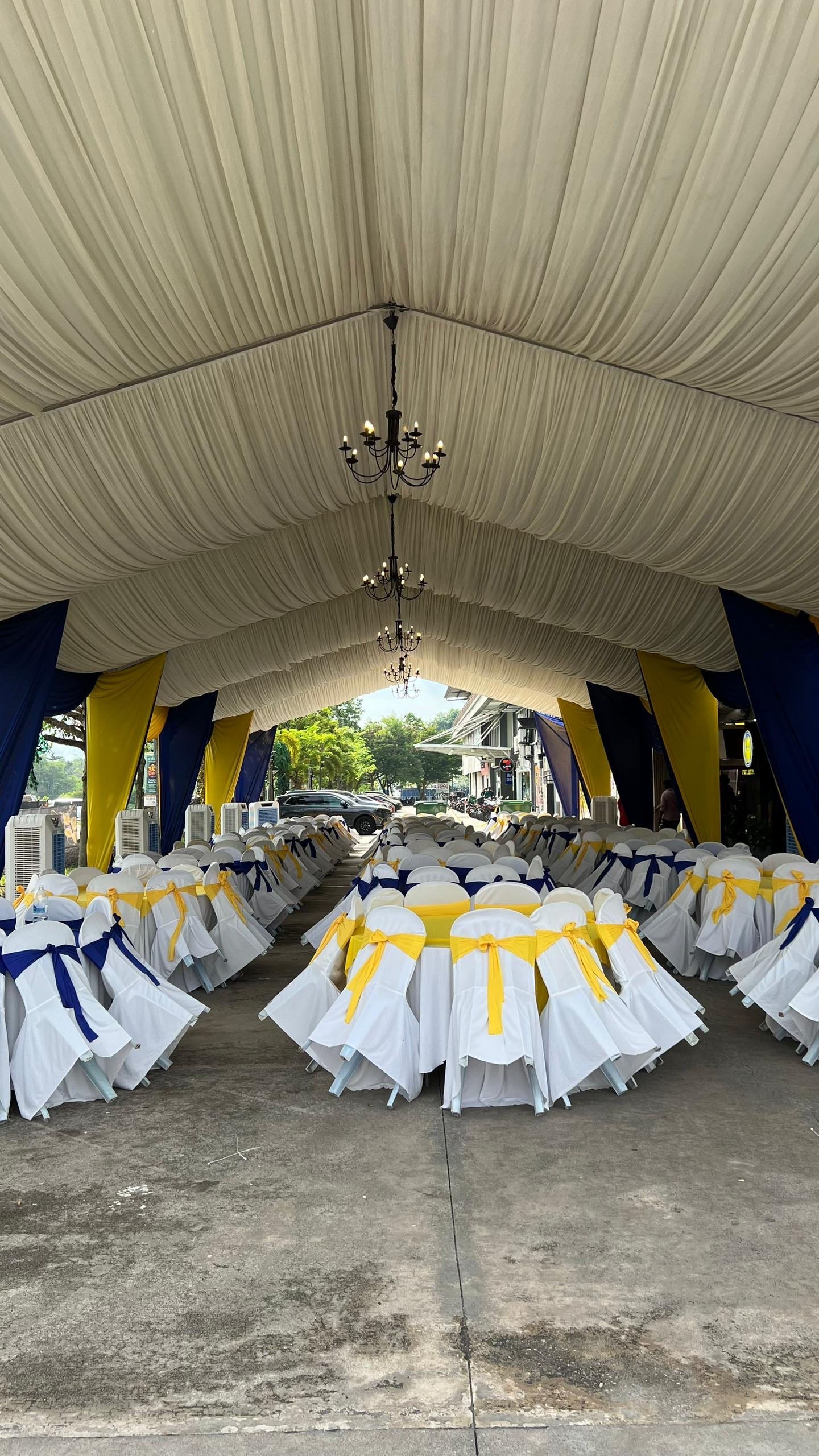 Event venue with round tables and chairs covered in white fabric, decorated with yellow and navy blue ribbons, under a large cream-colored fabric ceiling with chandeliers, chairs aligned in rows, and parked cars visible outside.