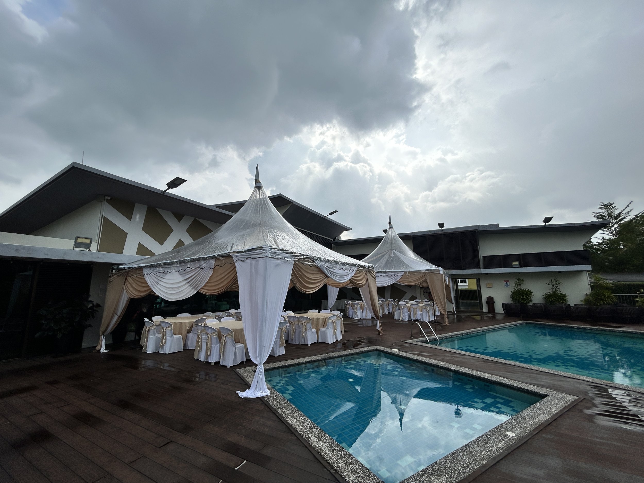 An outdoor poolside area with two white tents decorated with draped fabric, tables, and chairs, set against a modern building with a pool in the foreground and a cloudy sky overhead.