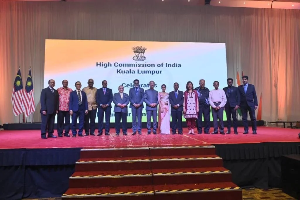 Group of people standing on a stage in front of a large screen displaying the text "High Commission of India, Kuala Lumpur, Celebrates" with the Indian flag symbols on the left side.