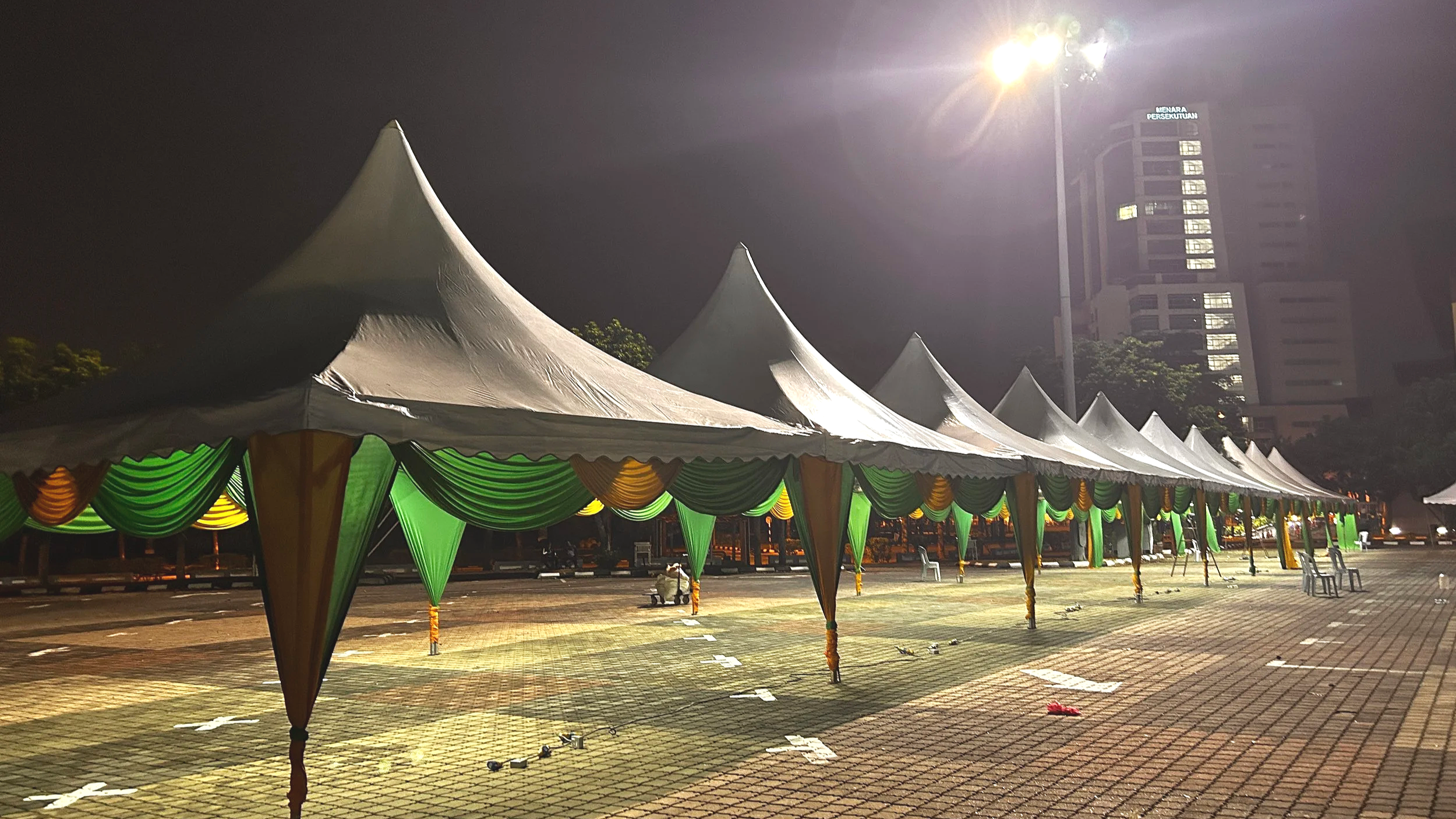 Nighttime scene showing large white tents with green and yellow drapes, set up in a parking lot or open area near tall buildings, illuminated by bright streetlights.