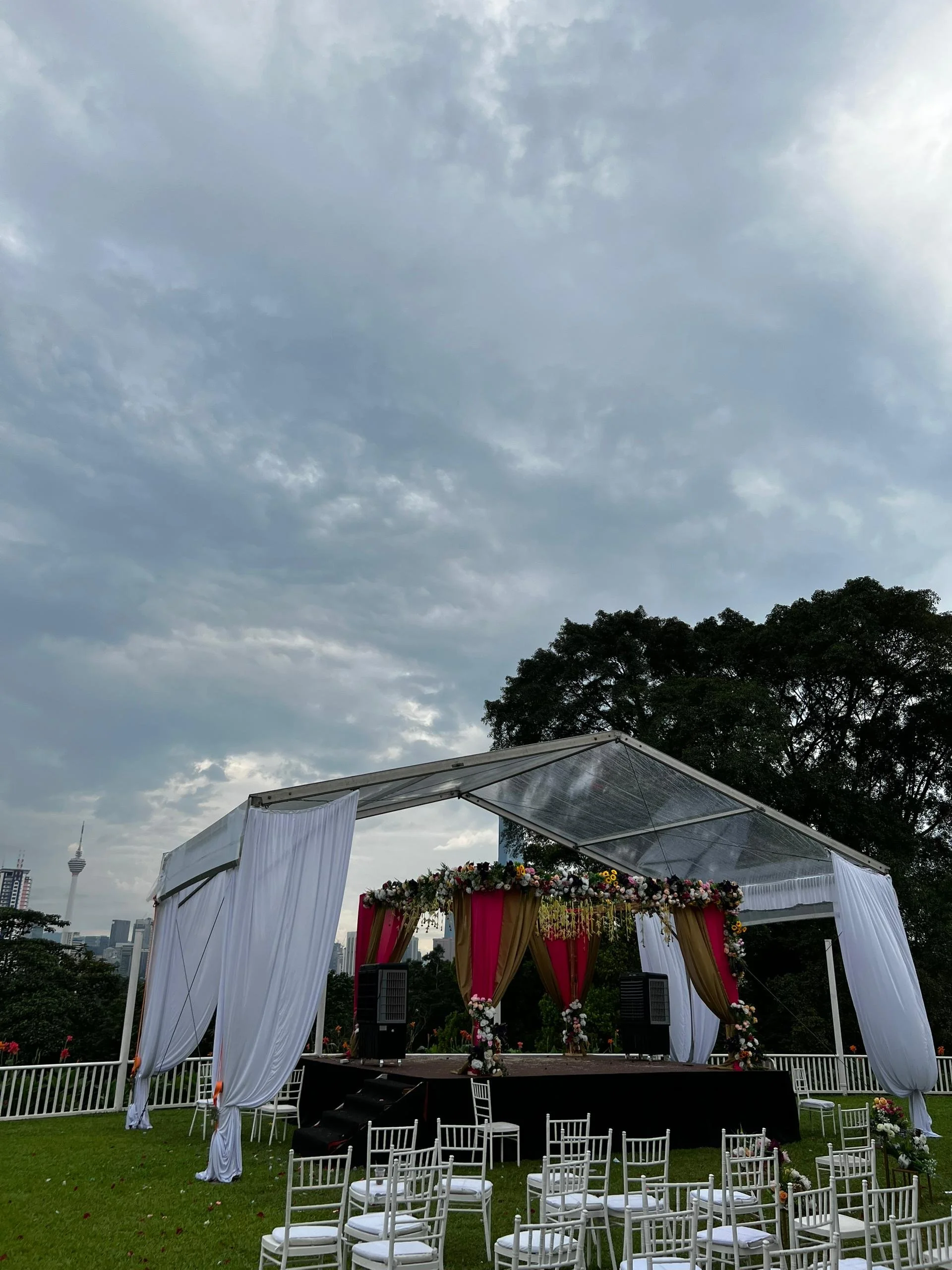 An outdoor wedding stage with a canopy decorated with flowers, surrounded by white chairs, and a city skyline with a tower visible in the background.