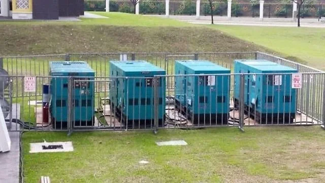 Four blue portable toilets are enclosed by a metal fence on a grassy area.