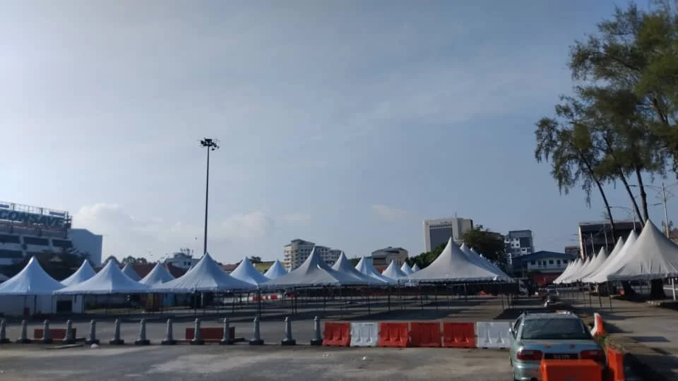 Rows of white tents set up in an open area, with some city buildings in the background, and a few cars and orange barriers in the foreground.