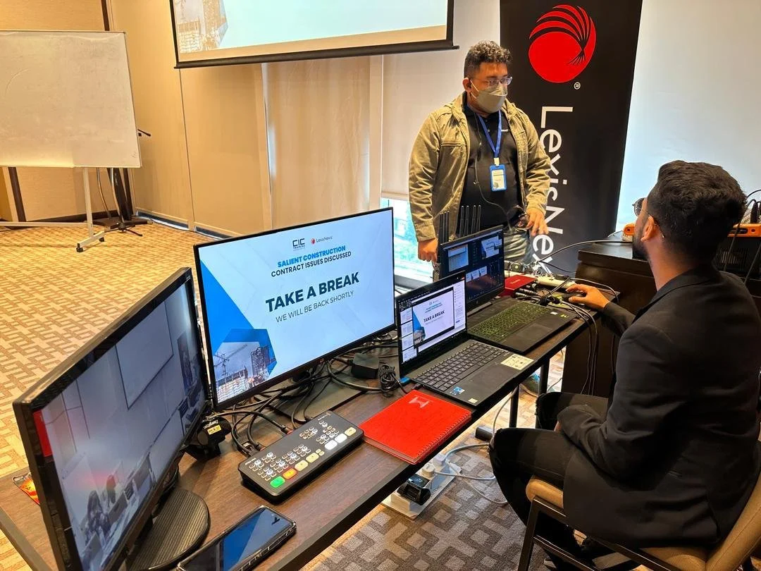 A man in a suit sitting at a table with multiple computers, screens, and electronic devices, in a conference room. There is a large monitor with a presentation slide that says "Take a break" and a man standing nearby, facing the seated man. A banner 