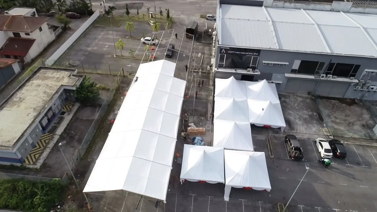 Aerial view of a parking lot with white tents set up for an outdoor event, adjacent to a commercial building, with a few parked cars and some trees in the background.