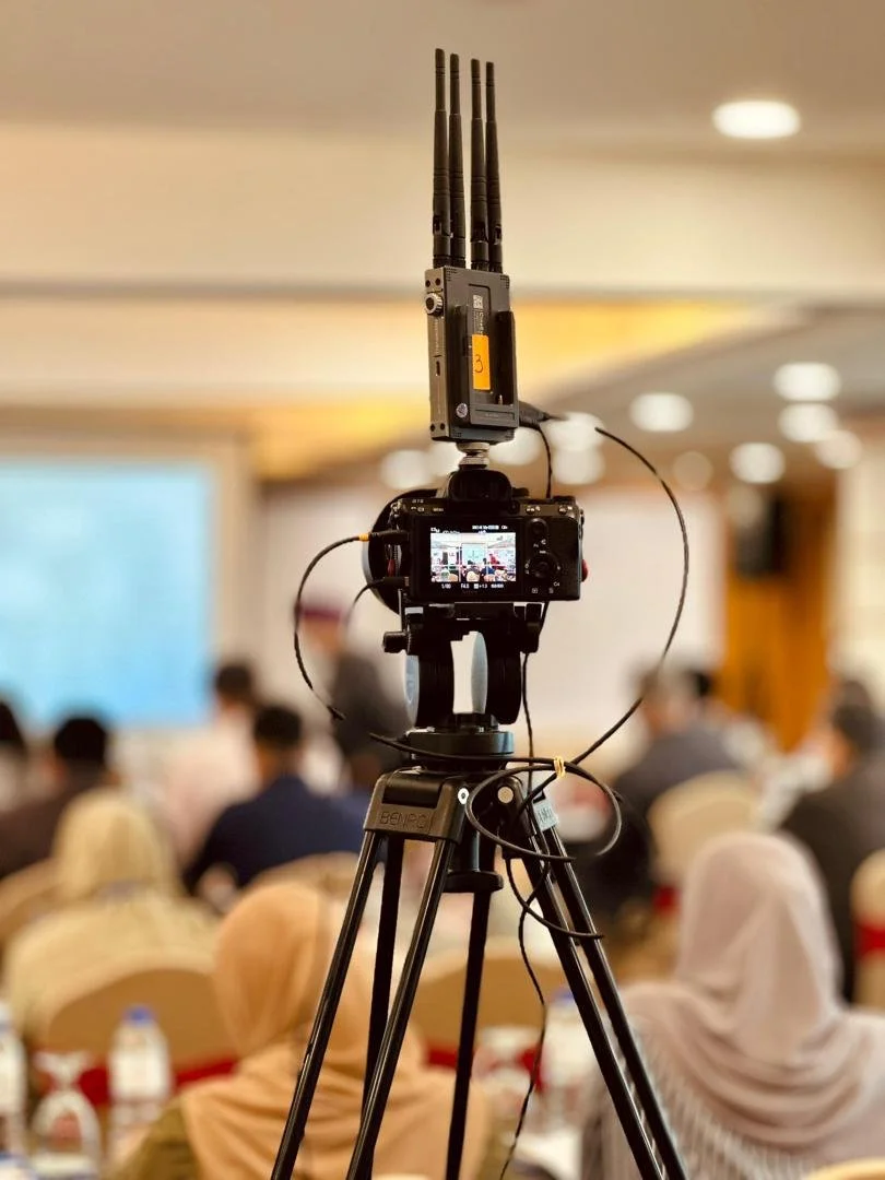 Camera mounted on tripod recording a conference with an audience in a well-lit room.