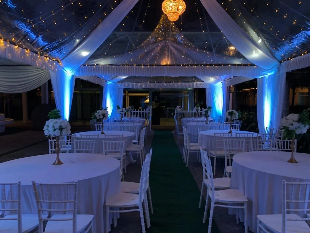 An elegant event space decorated with white drapes, string lights, and floral centerpieces on round tables. The ceiling features a chandelier and twinkling lights, with a green carpet aisle in the center.