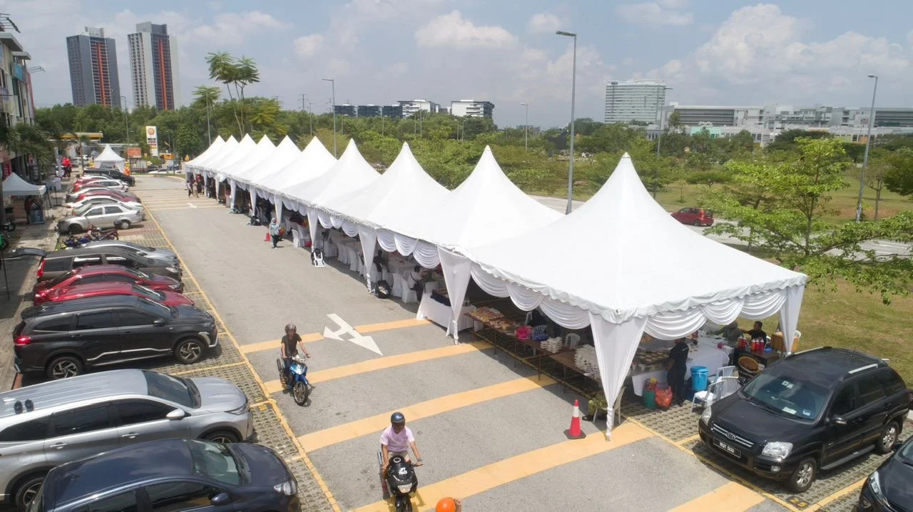 Outdoor market with white tents in a parking lot, cars parked along the sides, and people walking and riding motorcycles.