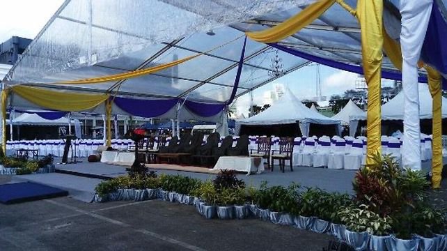 Empty outdoor event tent decorated with yellow and purple drapes, with tables and chairs set up underneath, surrounded by plants and flowers.