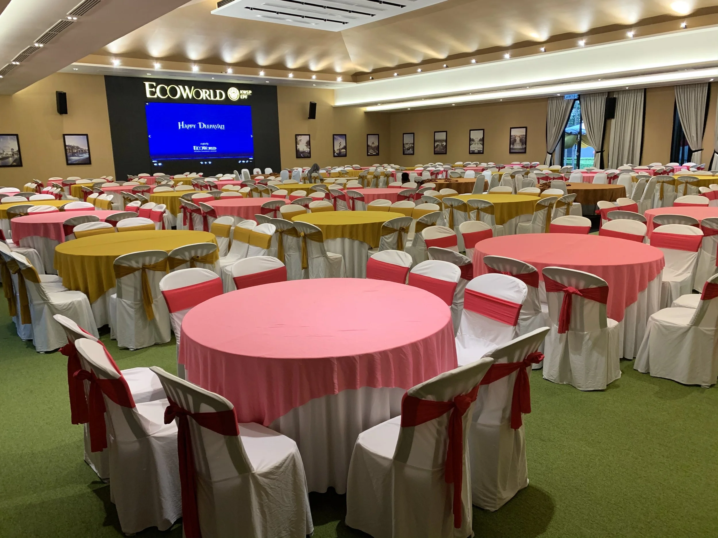 A large conference or banquet room with round tables covered in pink tablecloths and white chairs draped in white covers with gold sashes. The room has a high ceiling with warm lighting, a large screen at the front displaying a message, and is decora