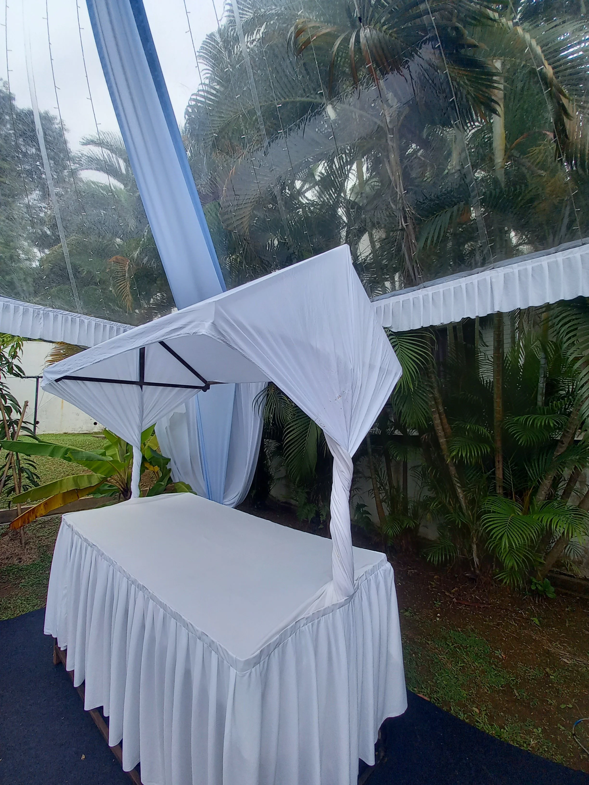 A white draped table with a white canopy in an outdoor setting surrounded by tropical plants and trees.