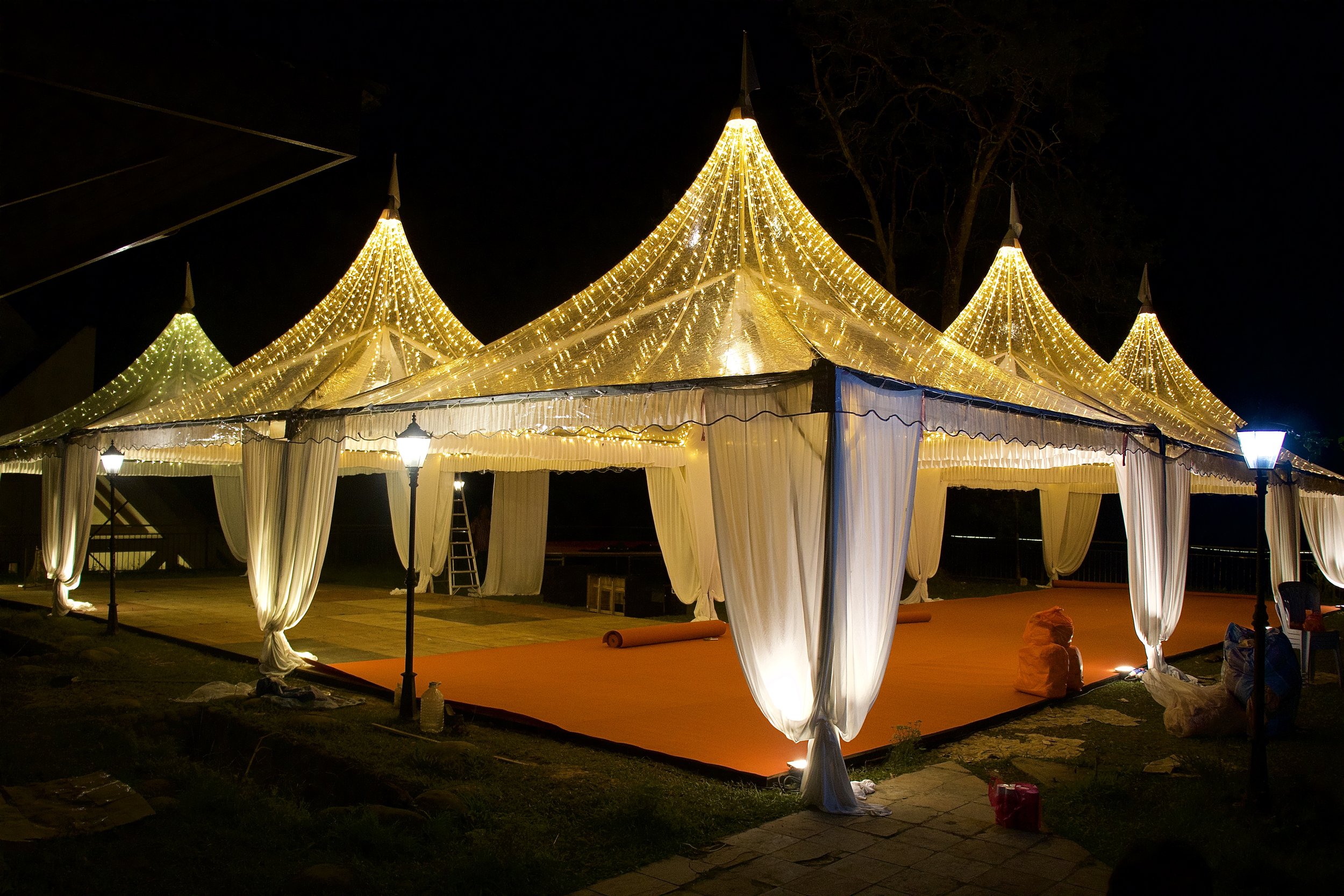 A large outdoor wedding tent illuminated with golden string lights at night, with draped white fabric walls and a red carpet underneath.