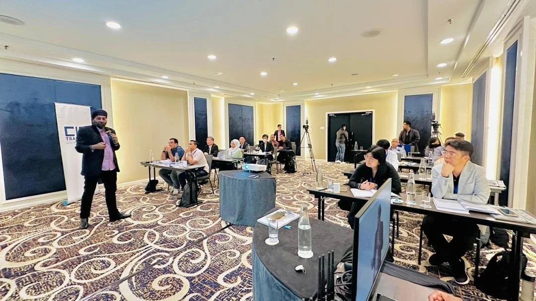 A man giving a presentation in a conference room with attendees seated at tables, some taking notes and listening.