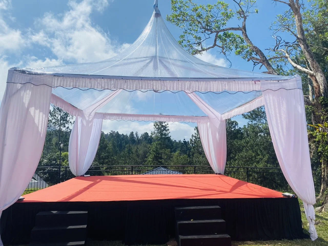 An outdoor stage with a canopy tent, pink curtains, and a red carpet, surrounded by trees and blue sky with clouds.