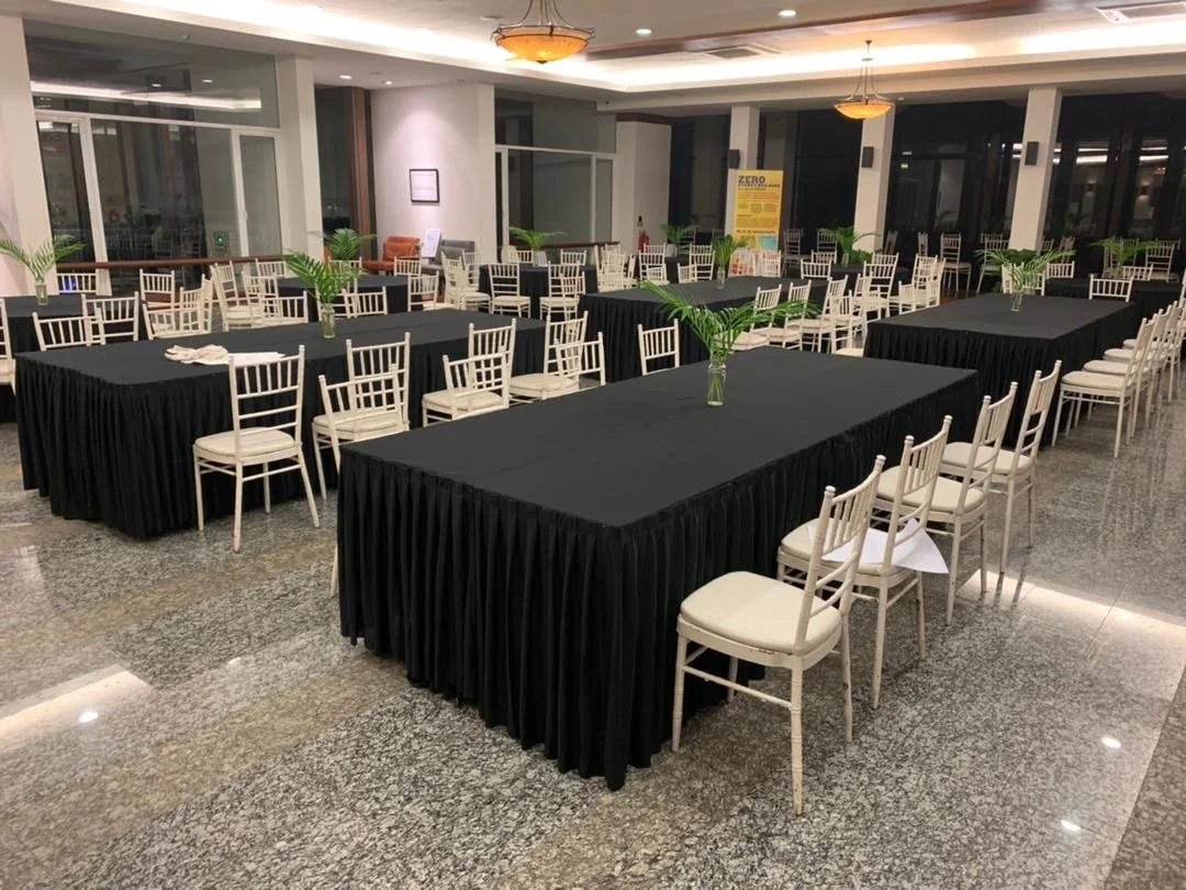 Conference room set up with long tables covered in black tablecloths, surrounded by white chairs, each table decorated with a small vase holding green palm leaves.