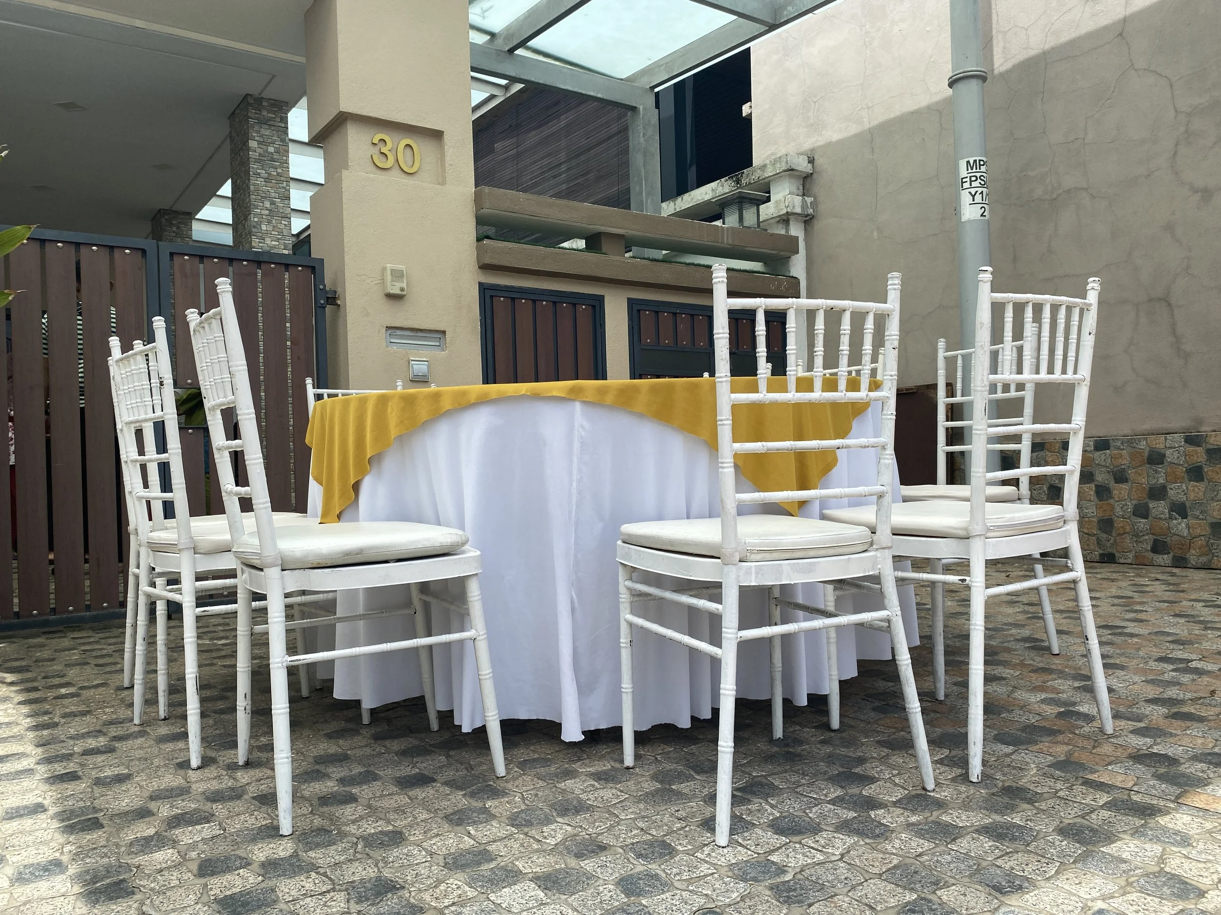 A round table with a white tablecloth and a yellow overlay is surrounded by white chairs, set outdoors on a cobblestone patio. In the background, there is a modern building with a beige wall, a brown wooden fence, and a gate, with a house number 30 o