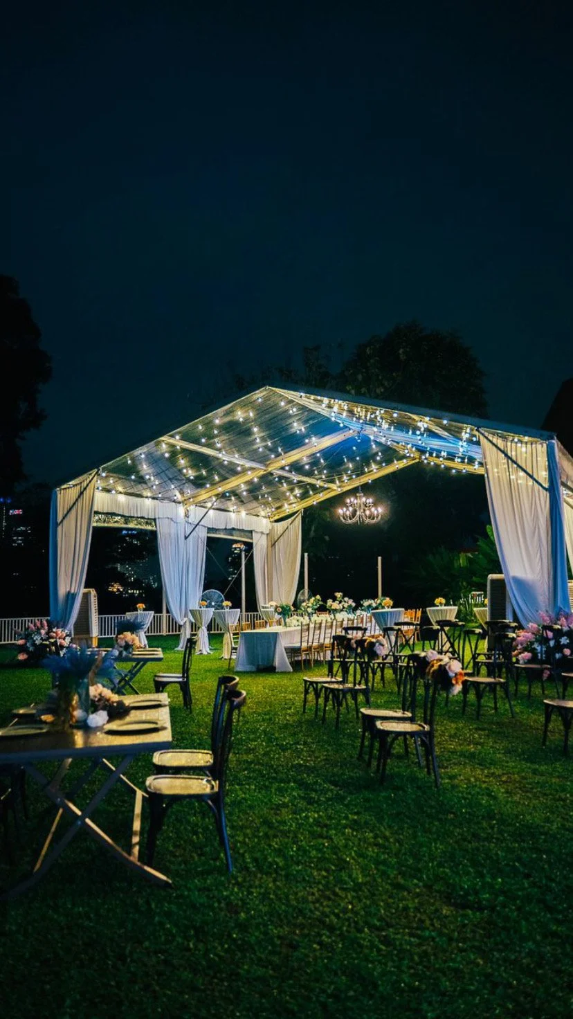 Outdoor event setup with tables, chairs, floral arrangements, a canopy with string lights, a chandelier, and a city skyline in the background at night.