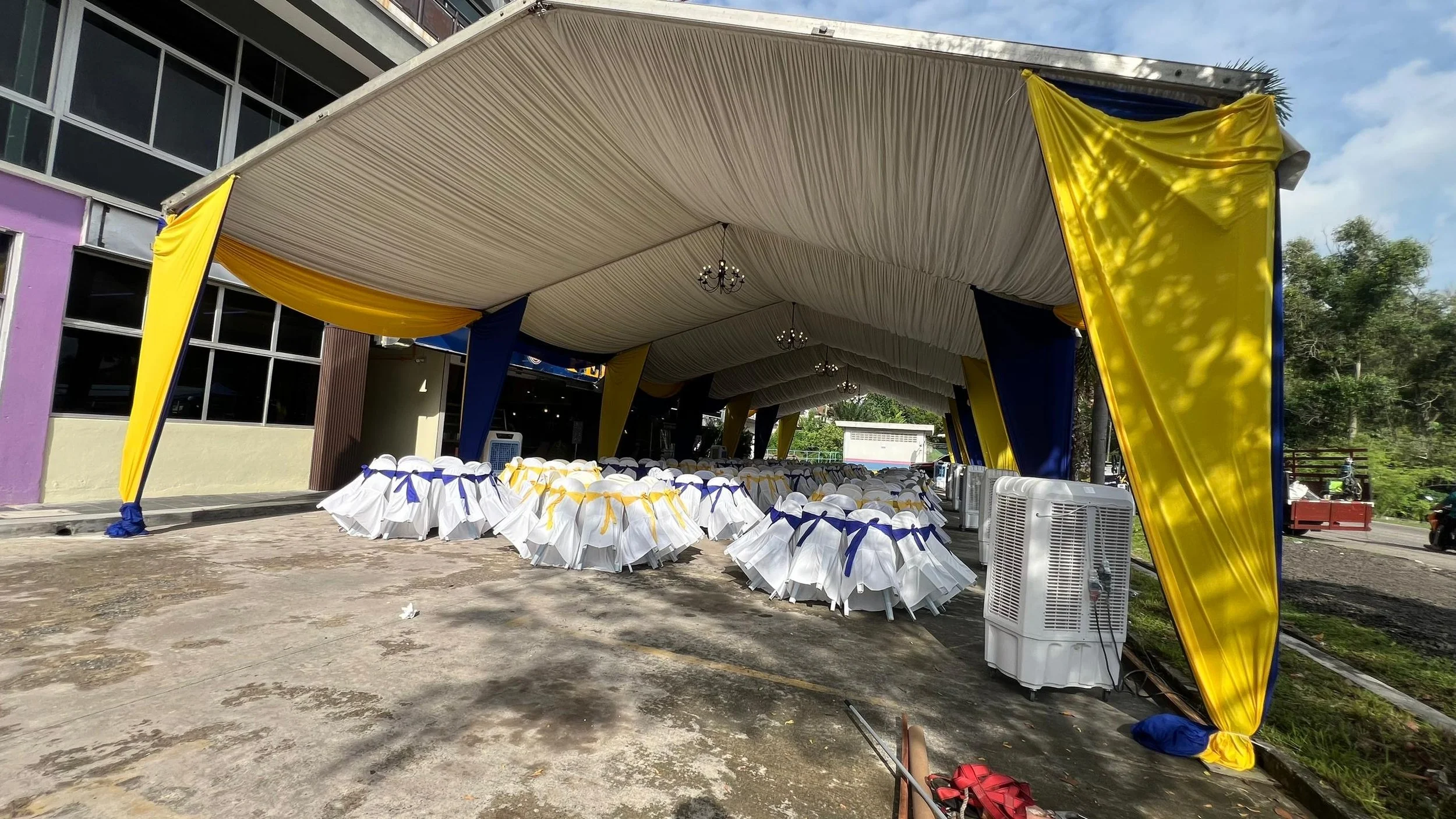 Setup for an outdoor event with tables and chairs covered in white cloth and decorated with blue and yellow ribbons, under a large beige canopy with yellow and blue drapes, located next to a building with purple and beige walls.