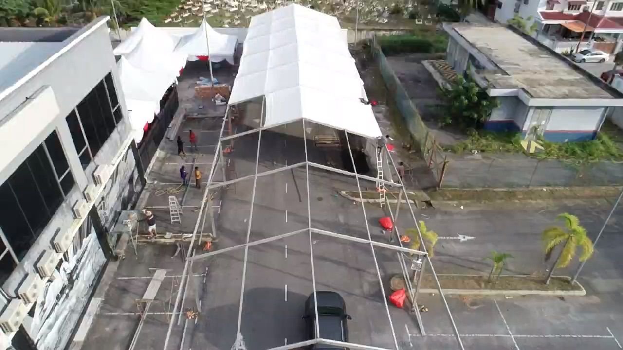 An aerial view shows a street with a temporary tent-like structure being assembled in front of a building. Several people are working on constructing the tent, with ladders and equipment around. The street is mostly empty, with a few cars parked and 