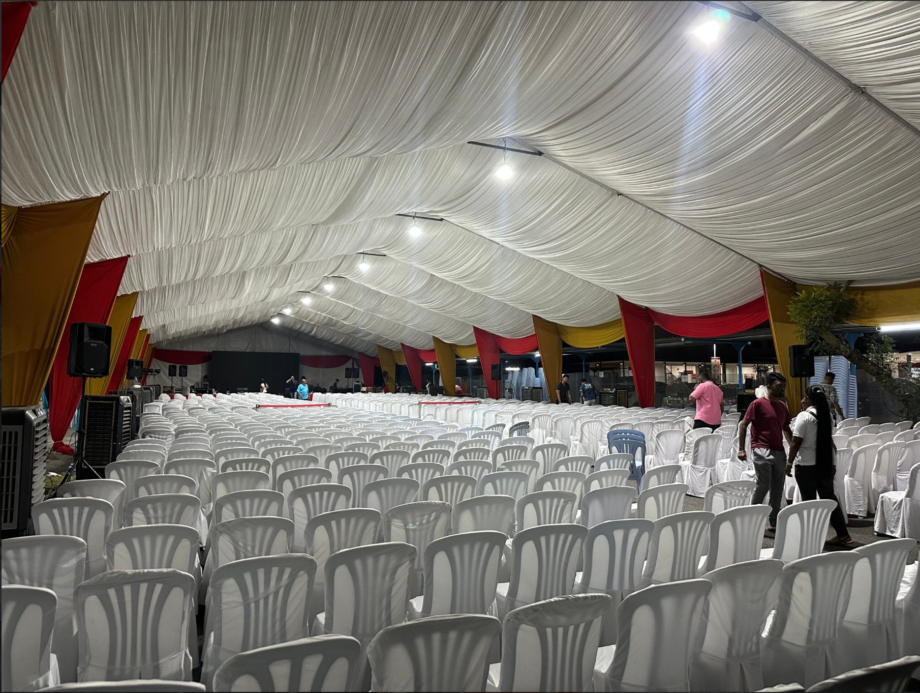 Large event tent interior with rows of white chairs facing a stage area, with people setting up and decorating for an event.