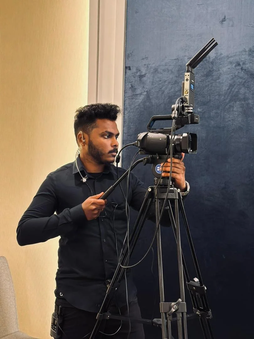 A man in black clothes operating a professional video camera on a tripod in an indoor setting.