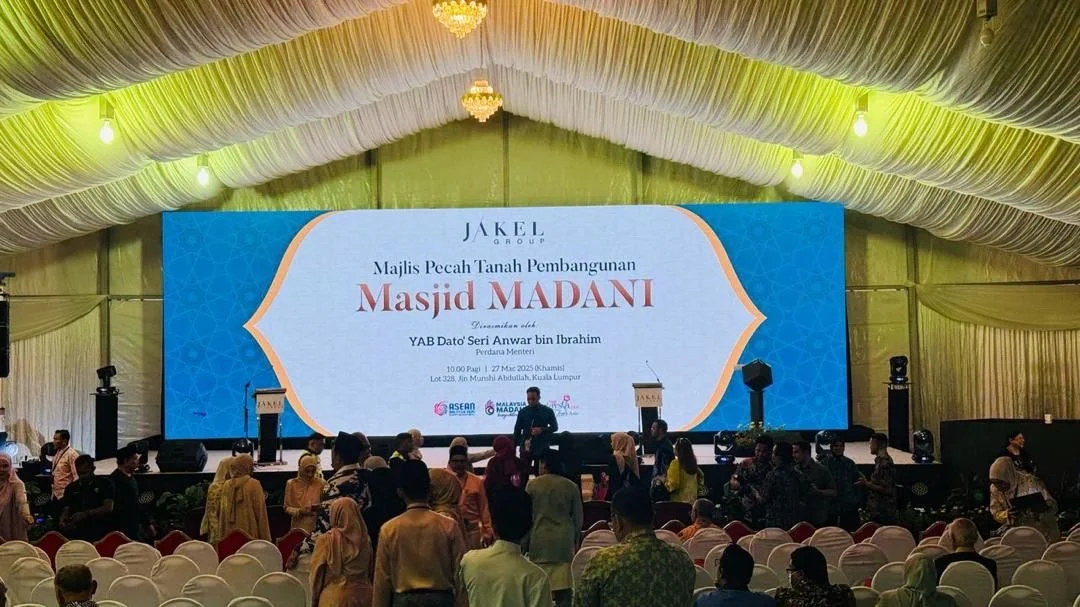 Large event hall with a stage and a big screen displaying a presentation in Malay language. The screen shows the event is related to the Masjid Madani construction project, held on May 27, 2023, in Kuala Lumpur. People are seated and standing in the 