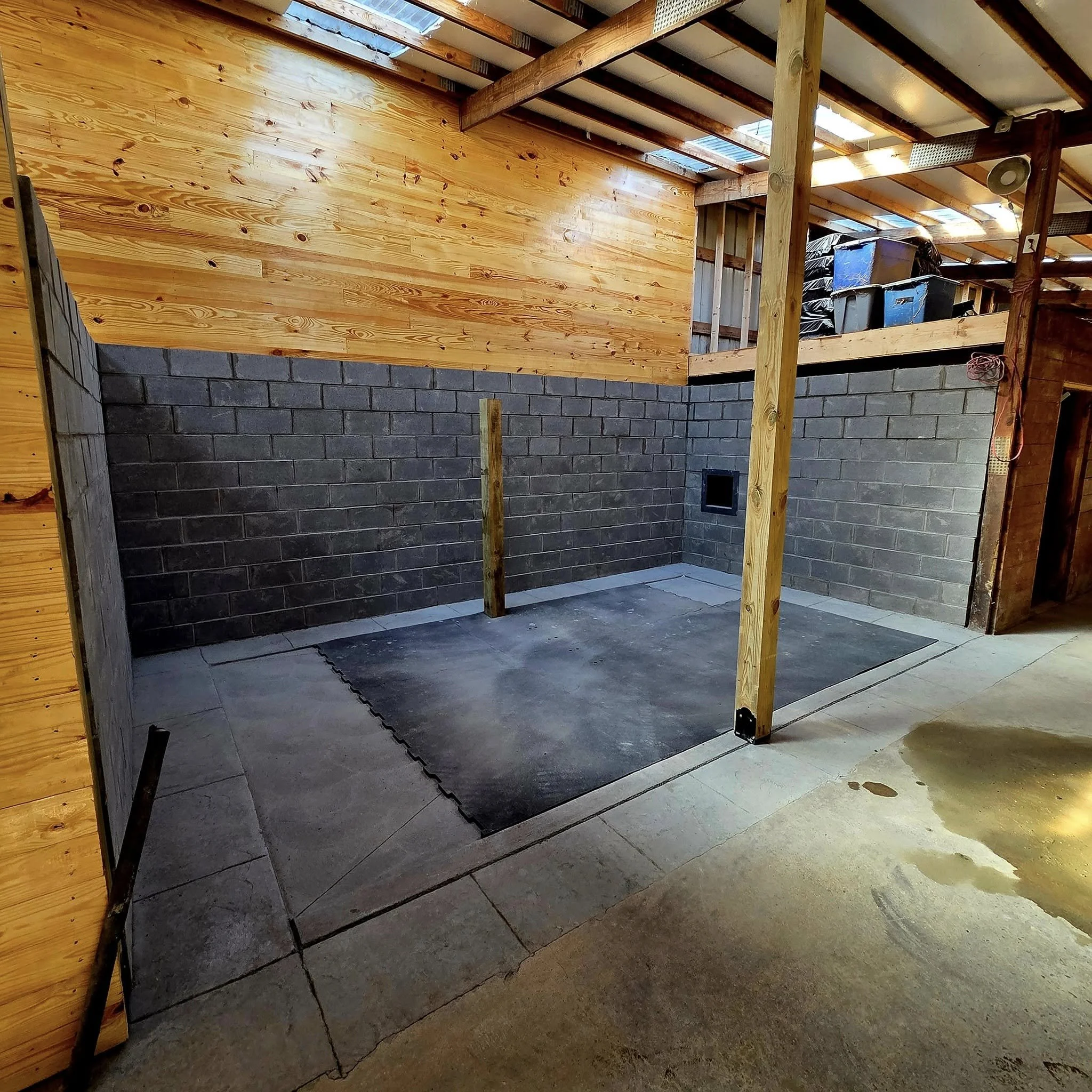 Interior of a building under construction with concrete block walls, wooden support beams, and a partially completed upper level. The floor area in the foreground has a rubber mat and some concrete, and a small window opening is visible on the wall.