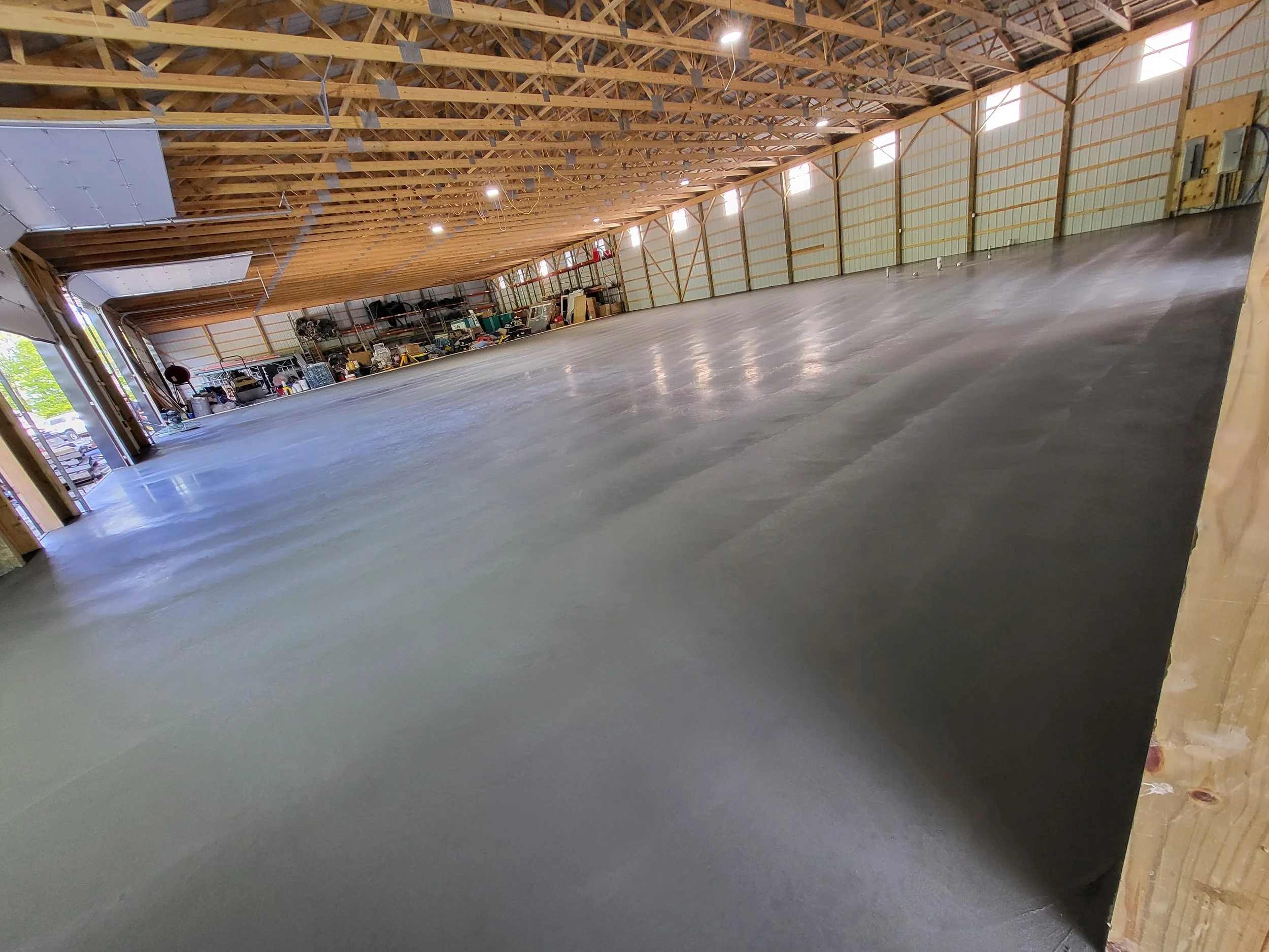 An interior of a large, empty warehouse with polished concrete floor and exposed wooden ceiling trusses. Shelving with tools and equipment at the back, open garage doors on the left side letting in natural light.