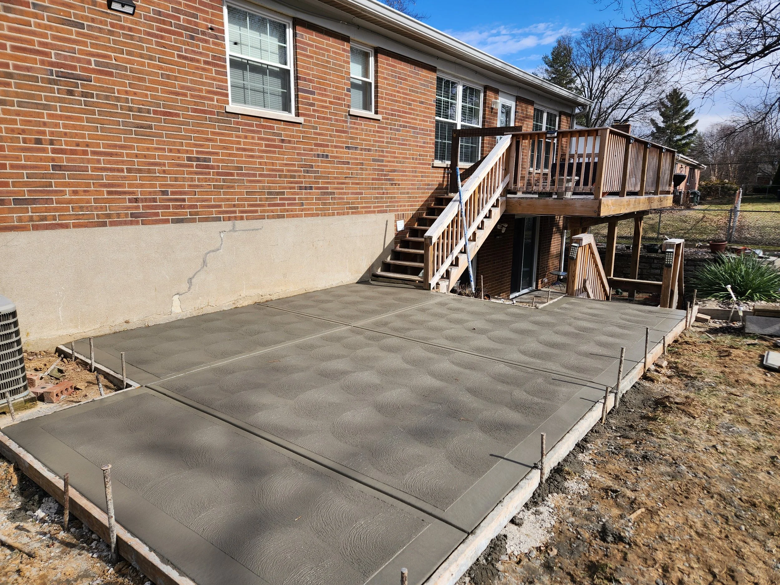 Freshly poured concrete patio in the backyard of a brick house, with a wooden staircase leading up to a raised deck.