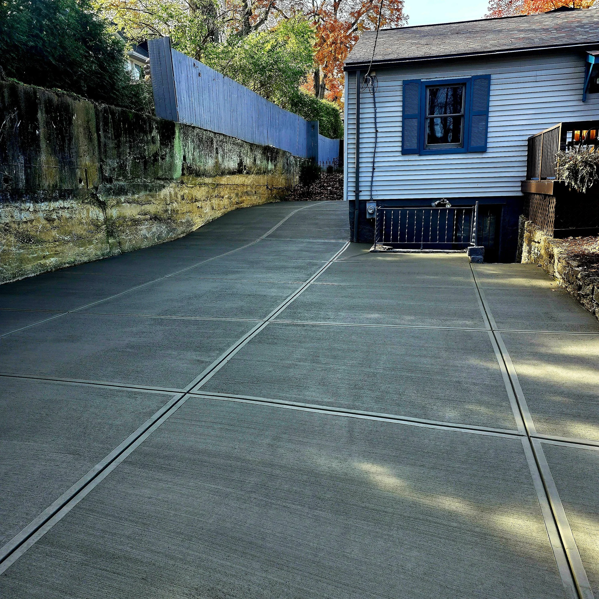 A residential driveway with concrete slabs and metal joints, adjacent to a house with blue shutters, on a sunny day with autumn trees in the background.