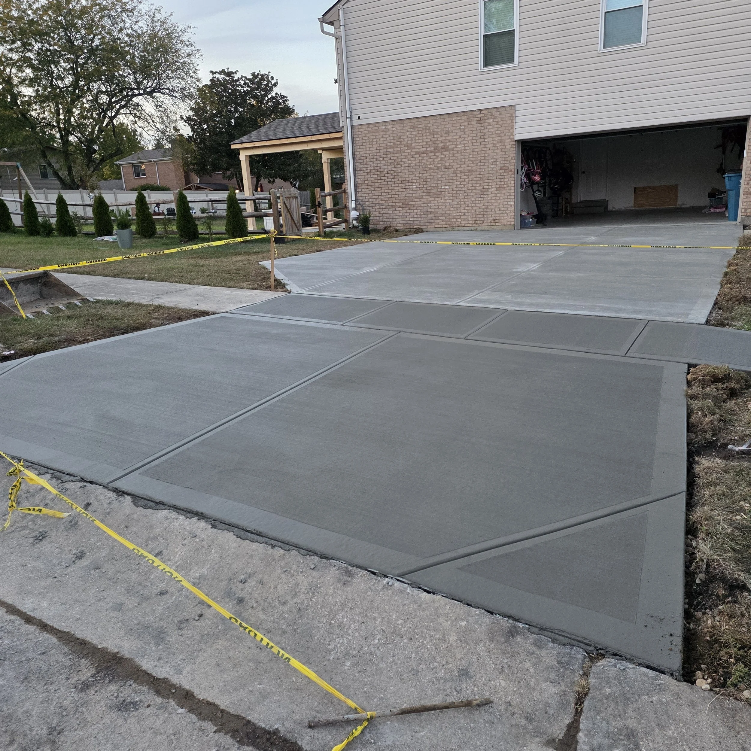 Freshly poured concrete driveway in front of a house, sectioned into separate slabs, with yellow caution tape around the construction area.