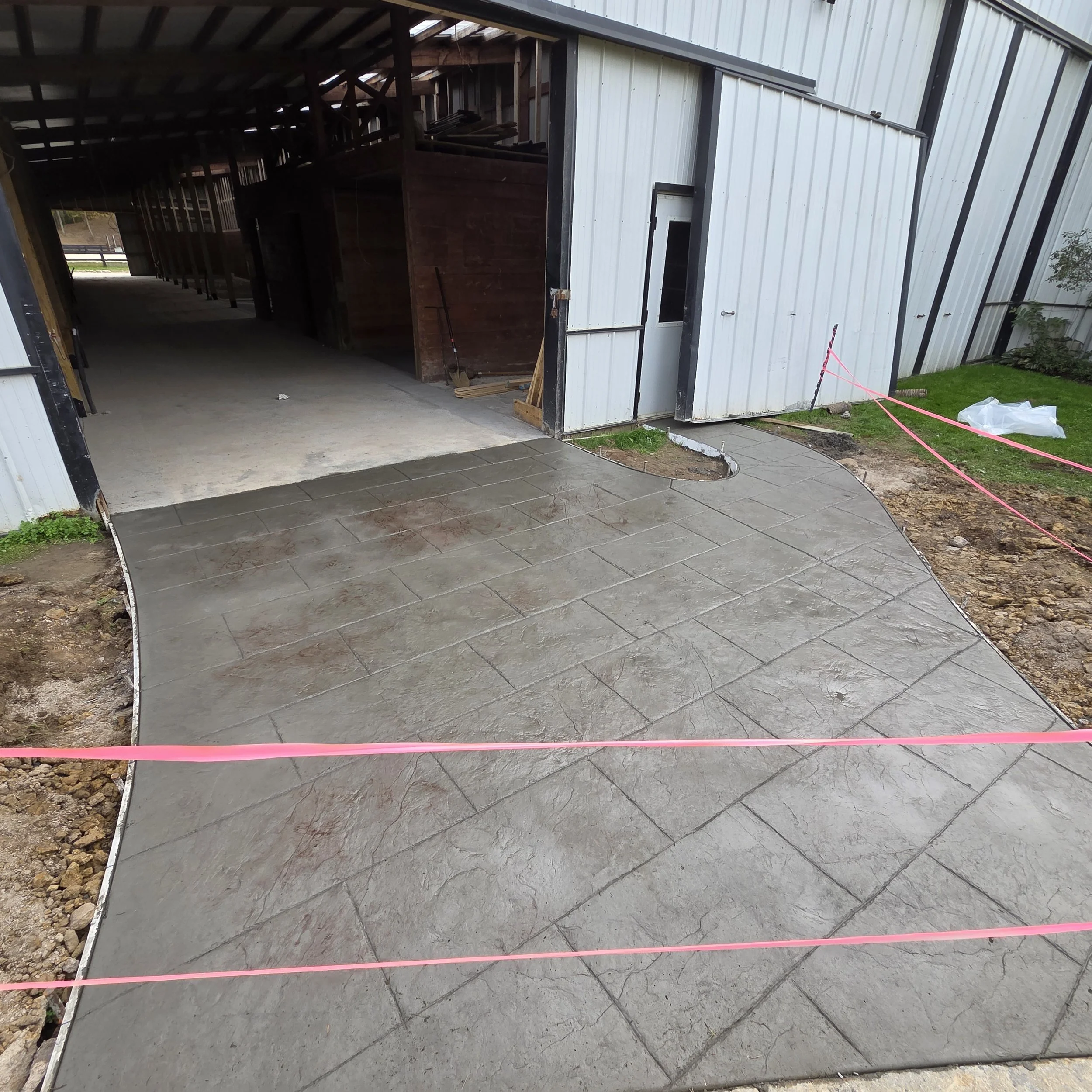 Construction site showing a newly poured concrete sidewalk in front of a building, with pink construction tape marking the perimeter.