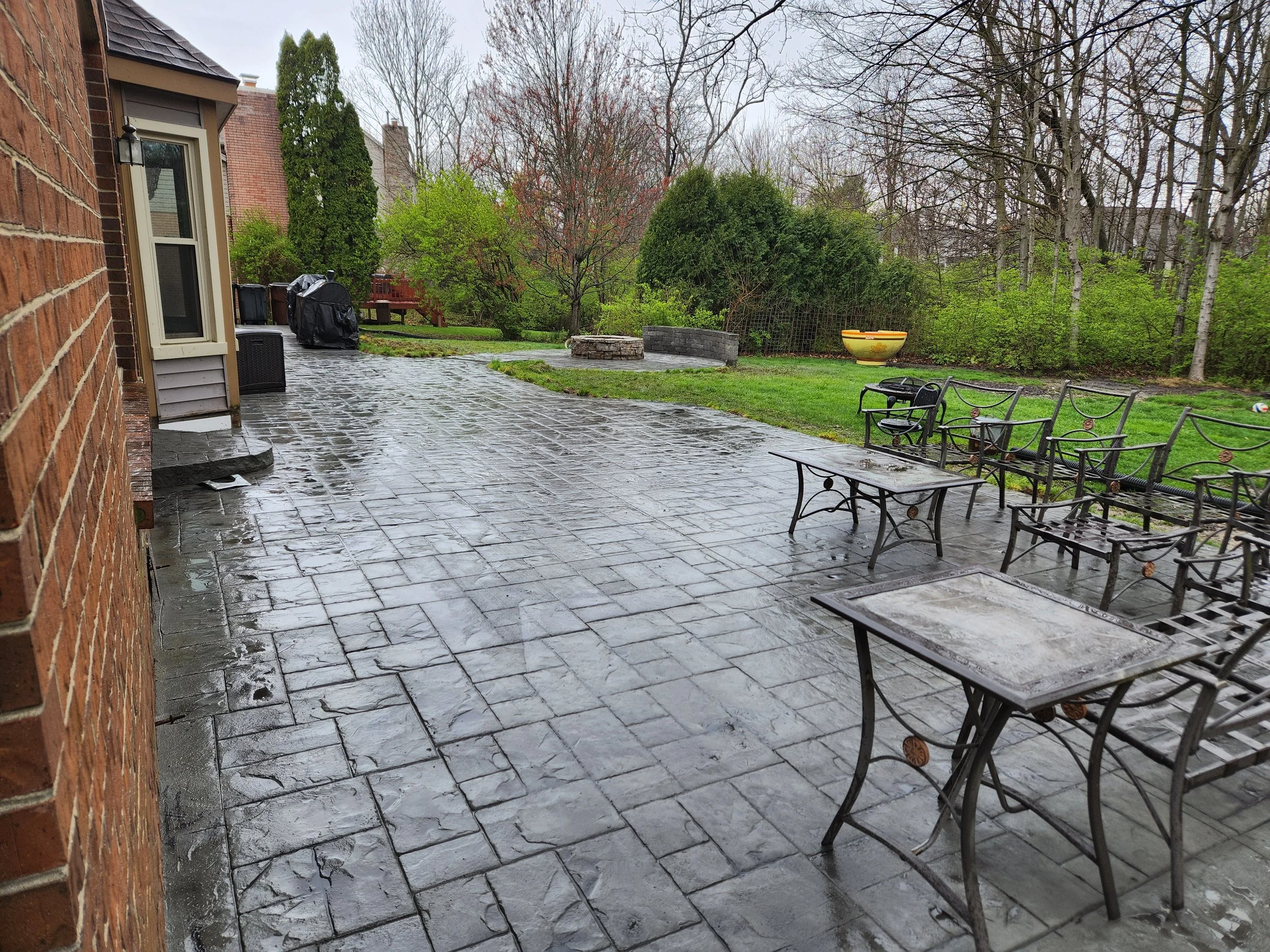 A wet outdoor patio area with a brick wall on the left side, several metal tables and chairs on the right, and greenery including trees and shrubs in the background on a rainy day.
