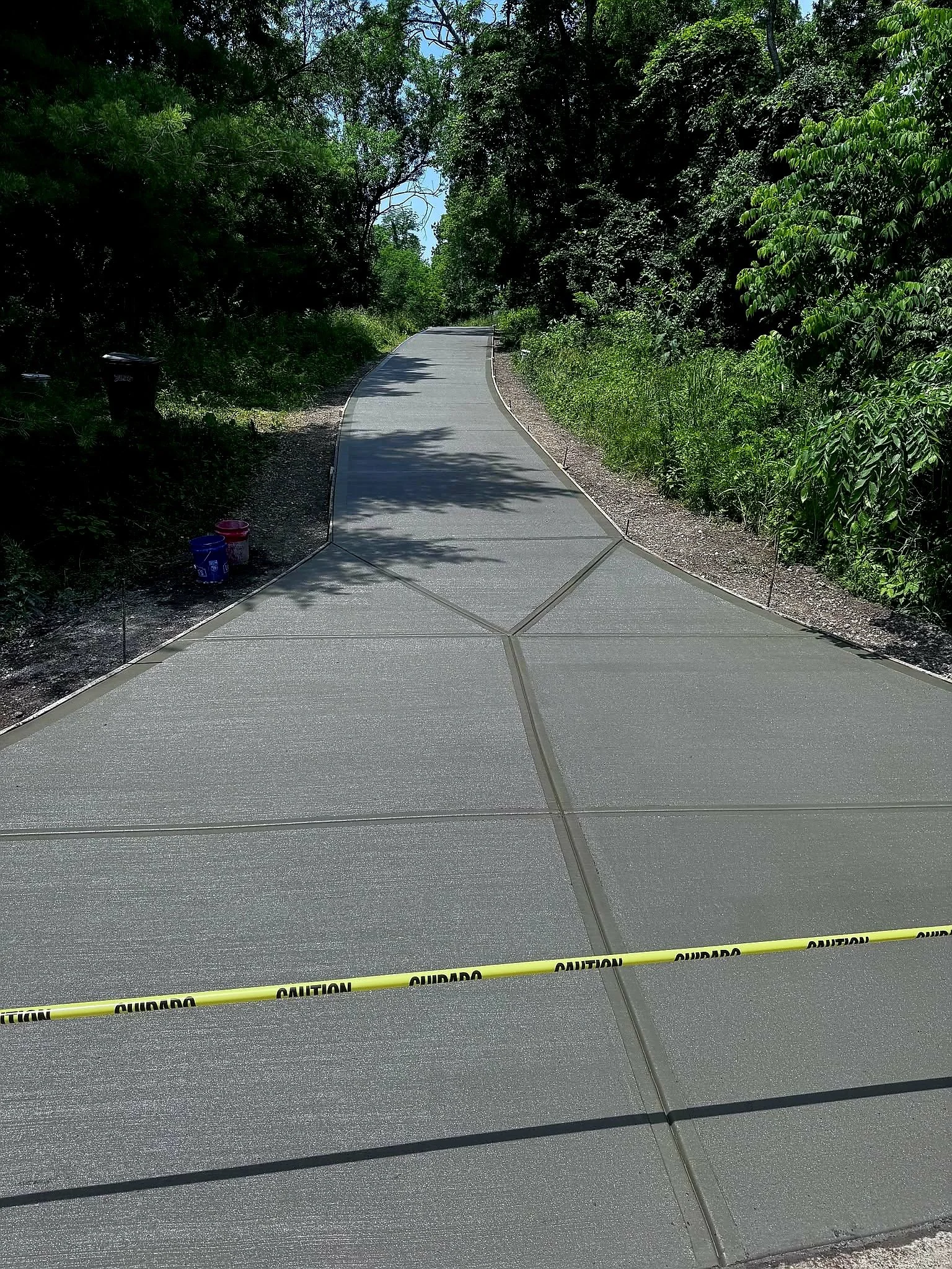A newly constructed concrete pathway with a caution tape across the front, surrounded by green trees and bushes on both sides, with a trash bin visible on the left side.