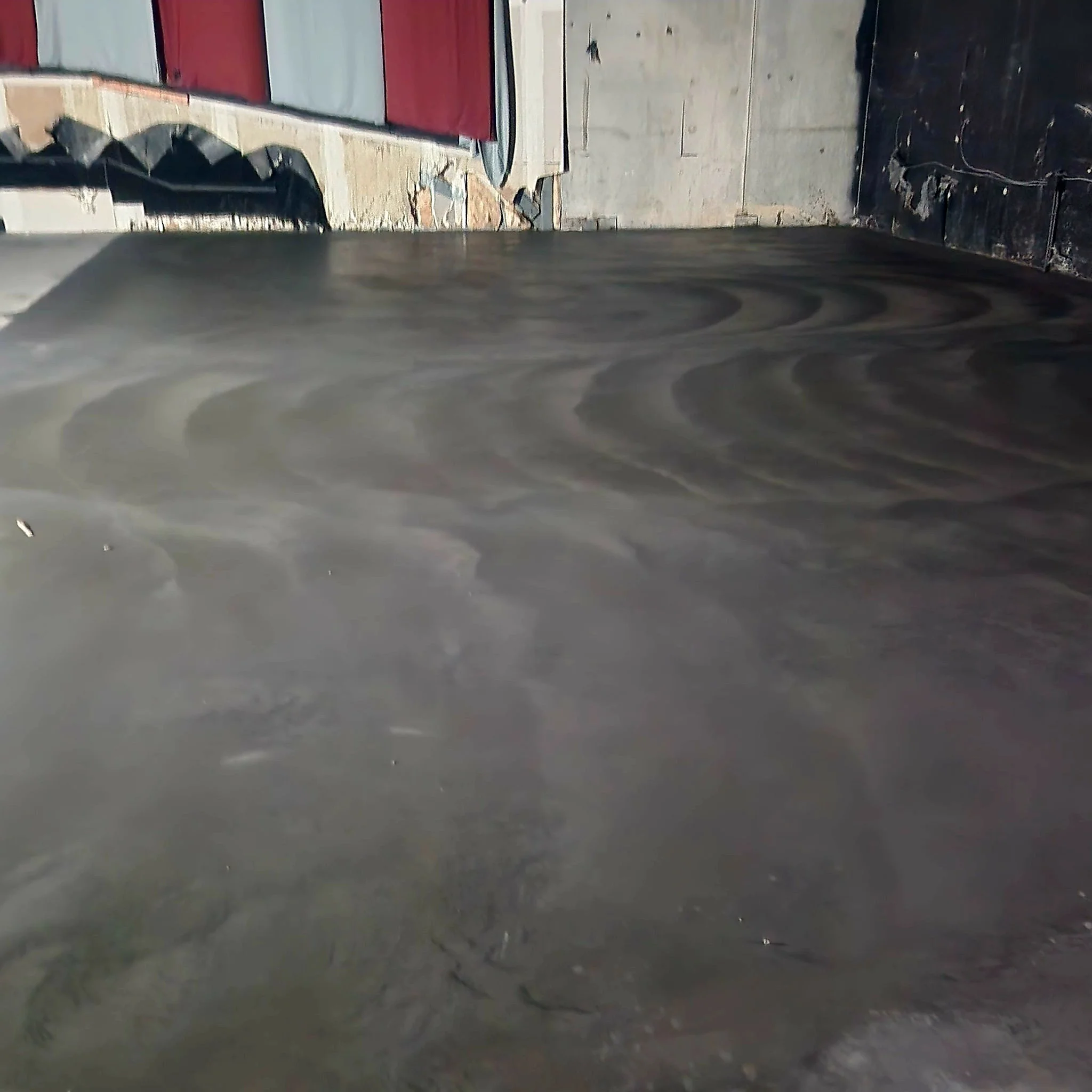 Close-up image of a wet concrete floor in an unfinished construction area with a brick wall, black pipes, and a grey concrete wall in the background.