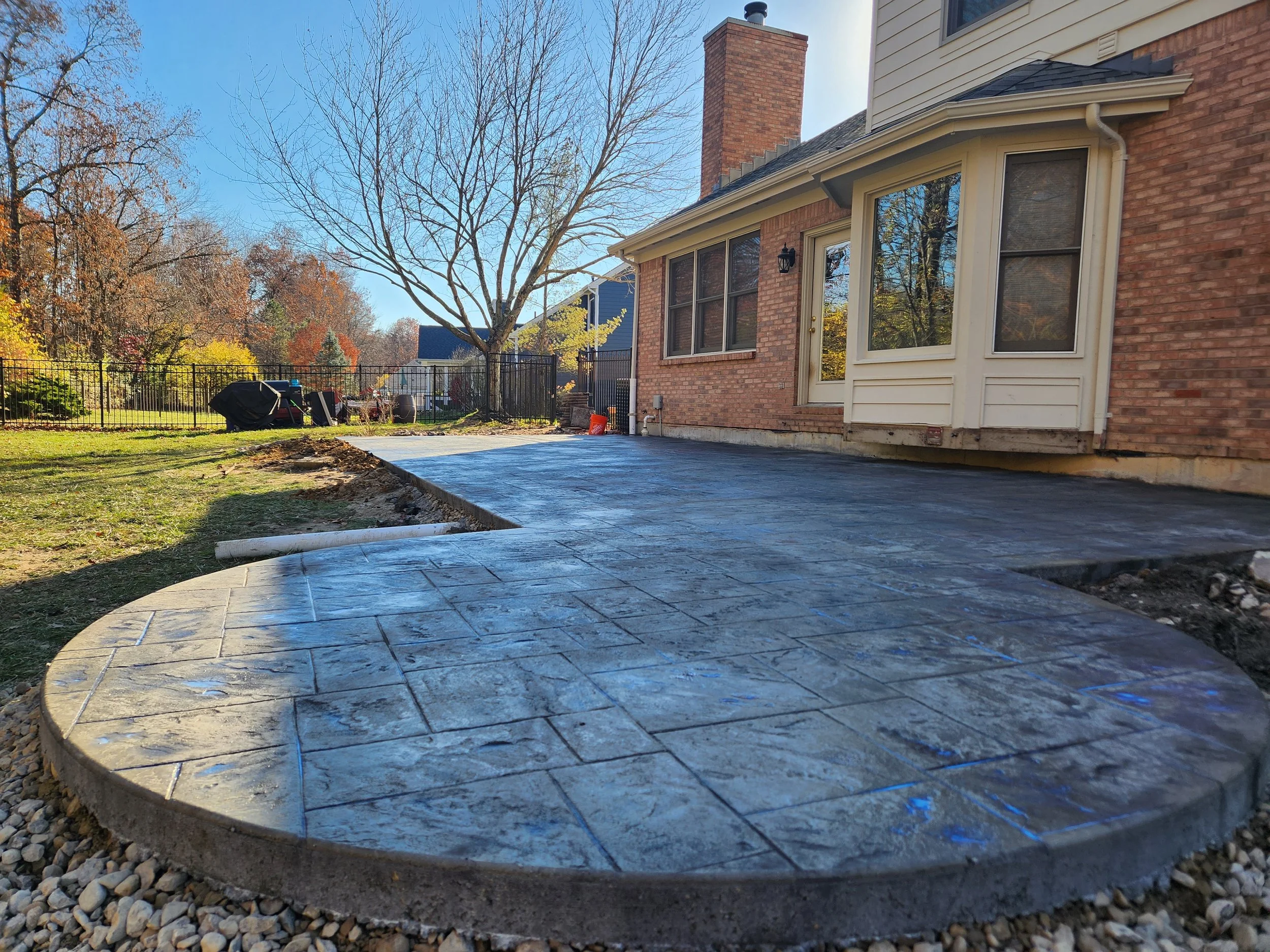 Newly poured concrete patio with stamped design attached to a brick house, in a backyard with a leafless tree, a black metal fence, and a clear blue sky.
