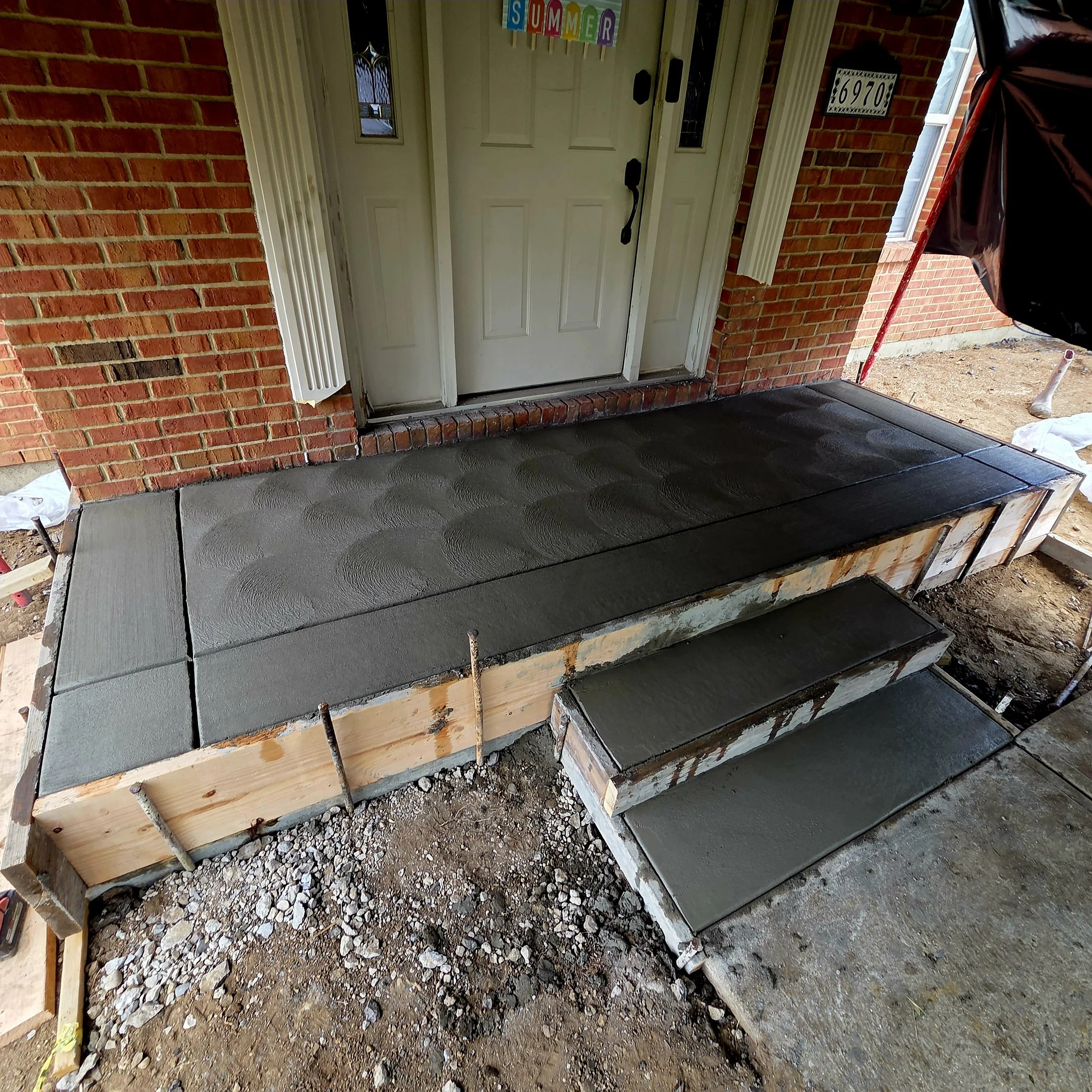 Concrete porch construction in progress in front of a house with a white door, red brick exterior, and colorful 'SUMMER' banner. New concrete steps and landing area are being poured, with some sections freshly smoothed and others still under work.