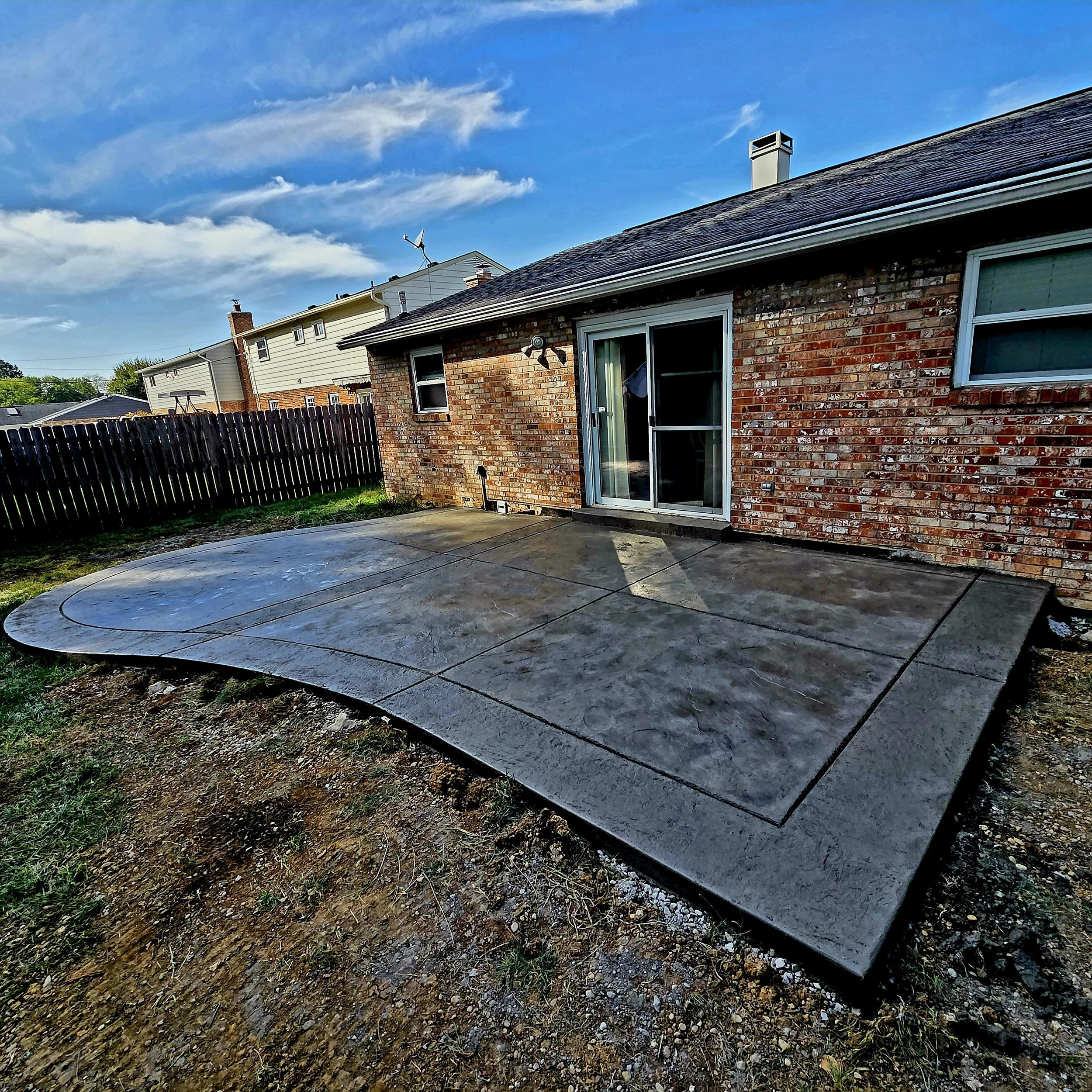 Backyard with a newly poured concrete patio, brick house with sliding glass door, wooden fence, and neighboring houses under a partly cloudy sky.