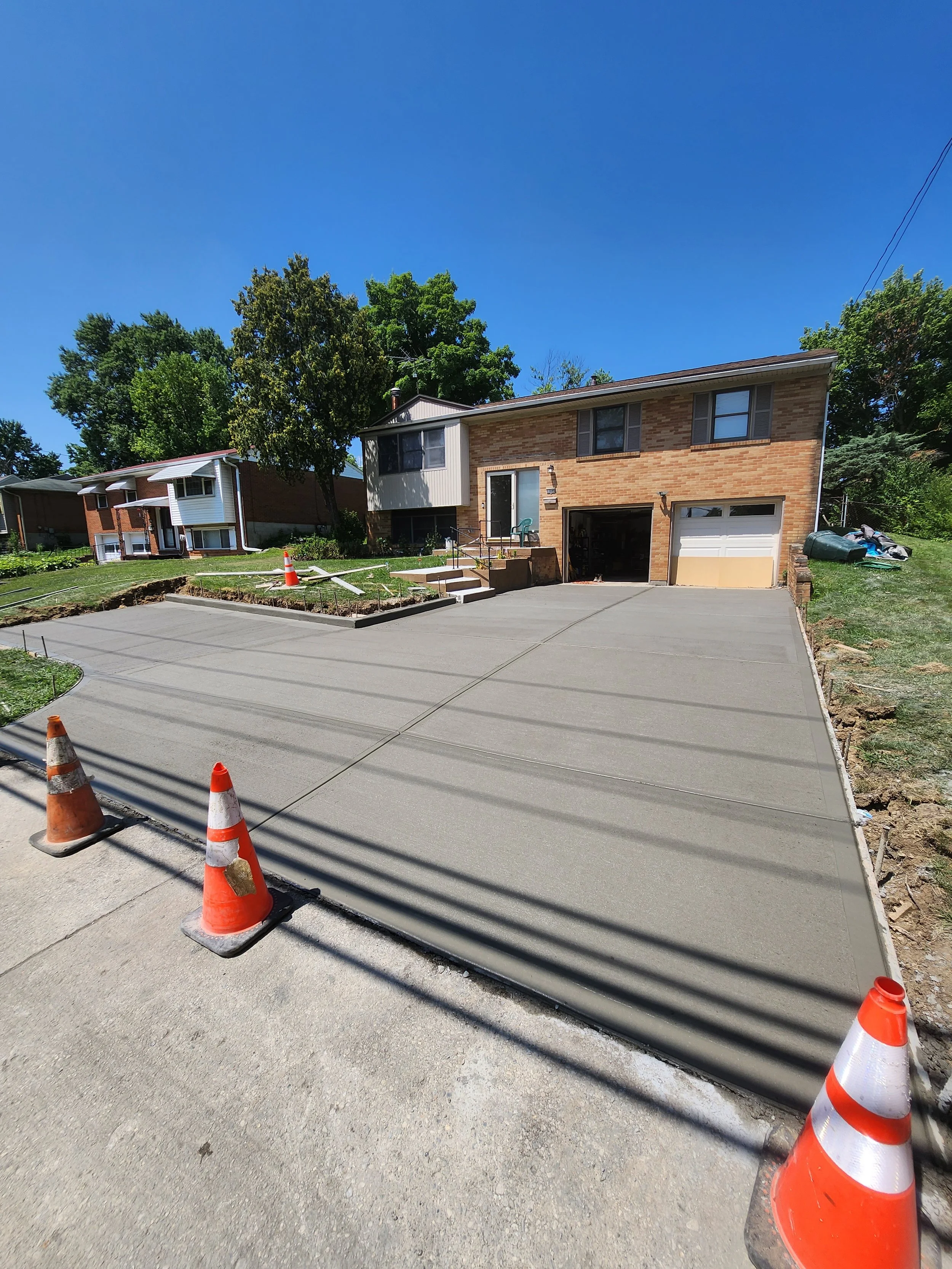 Newly poured concrete driveway with orange construction cones in front, leading to a brick house with an attached garage, on a sunny day with a blue sky and nearby trees.