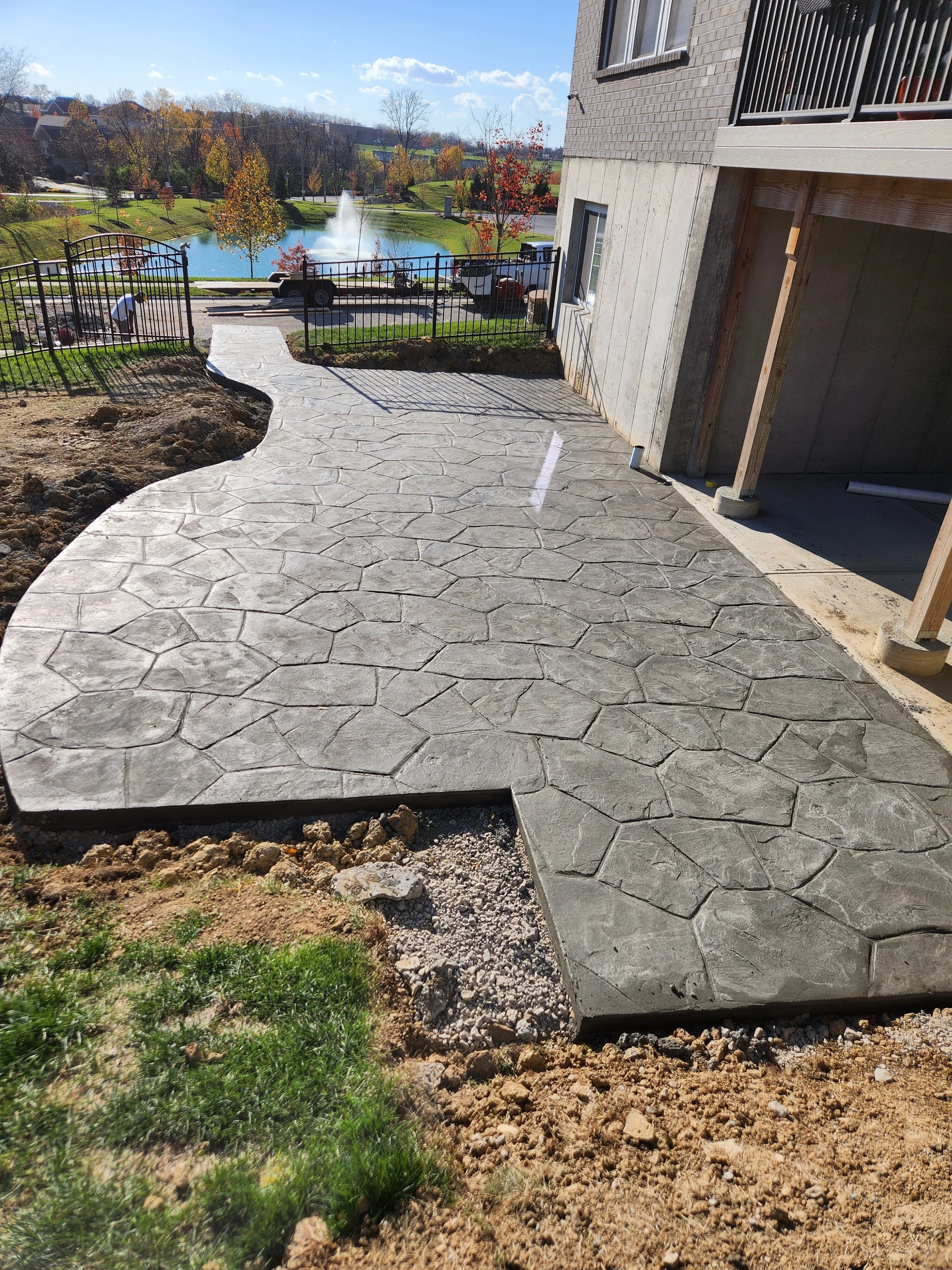 Newly poured concrete patio with a stamped stone pattern next to a house under construction, overlooking a park with a pond, fountain, trees, and parked cars.