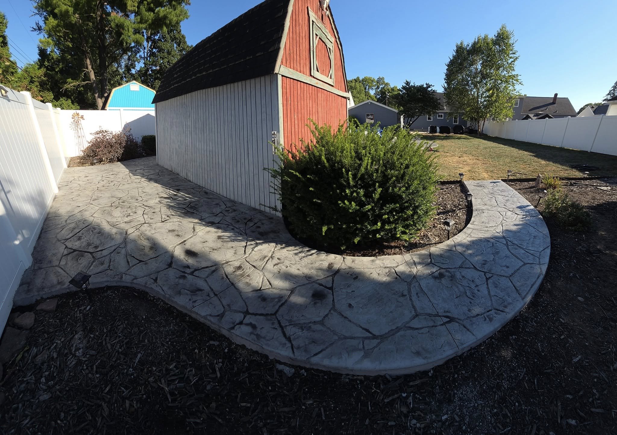A backyard with a concrete stamped pathway, a red barn-style shed, a green bush, a tree, and a white fence under a clear blue sky.