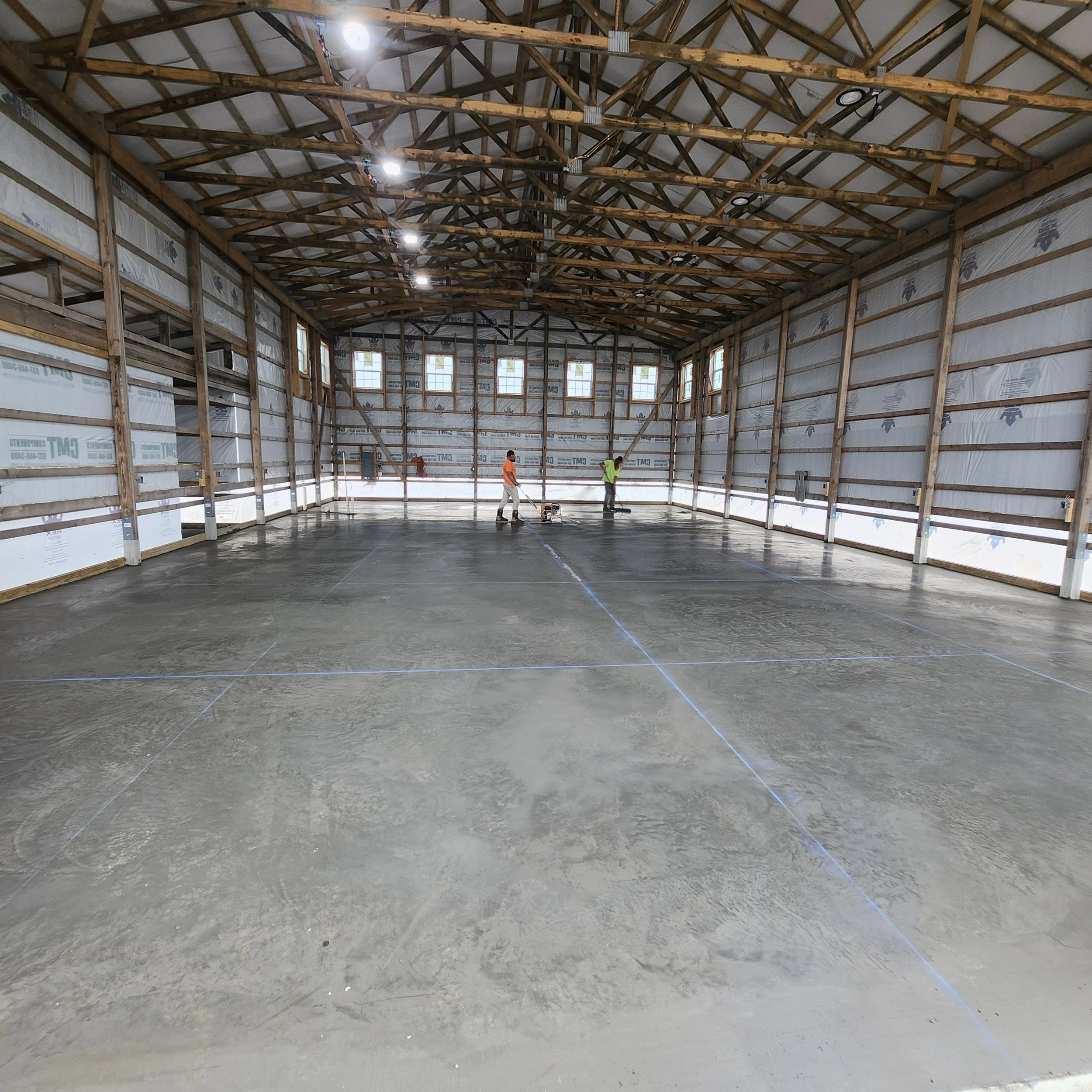 Inside a large wooden structure under construction, workers are leveling a concrete floor marked with blue chalk lines.