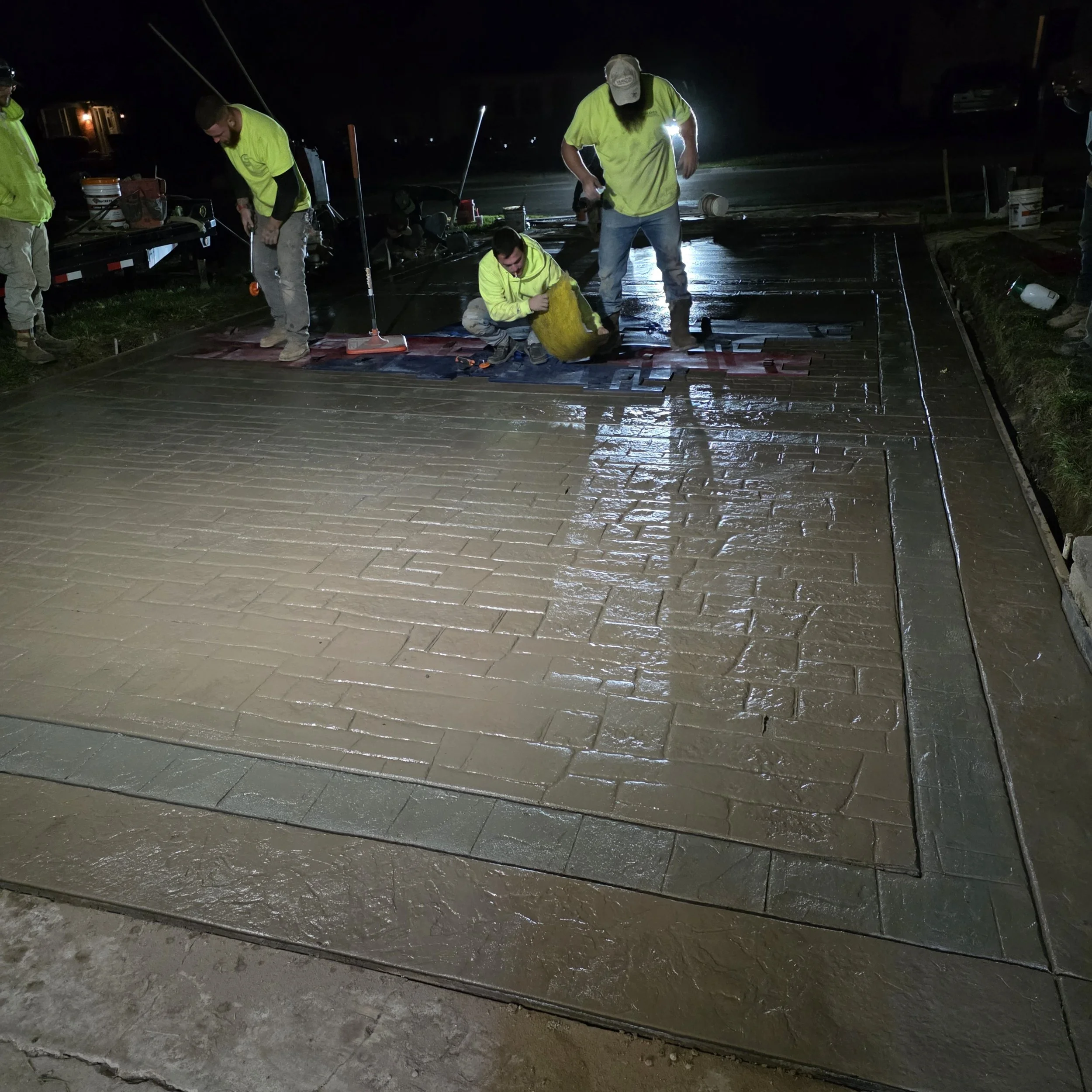 Workers installing decorative concrete paving stones at night, with construction tools and equipment visible.