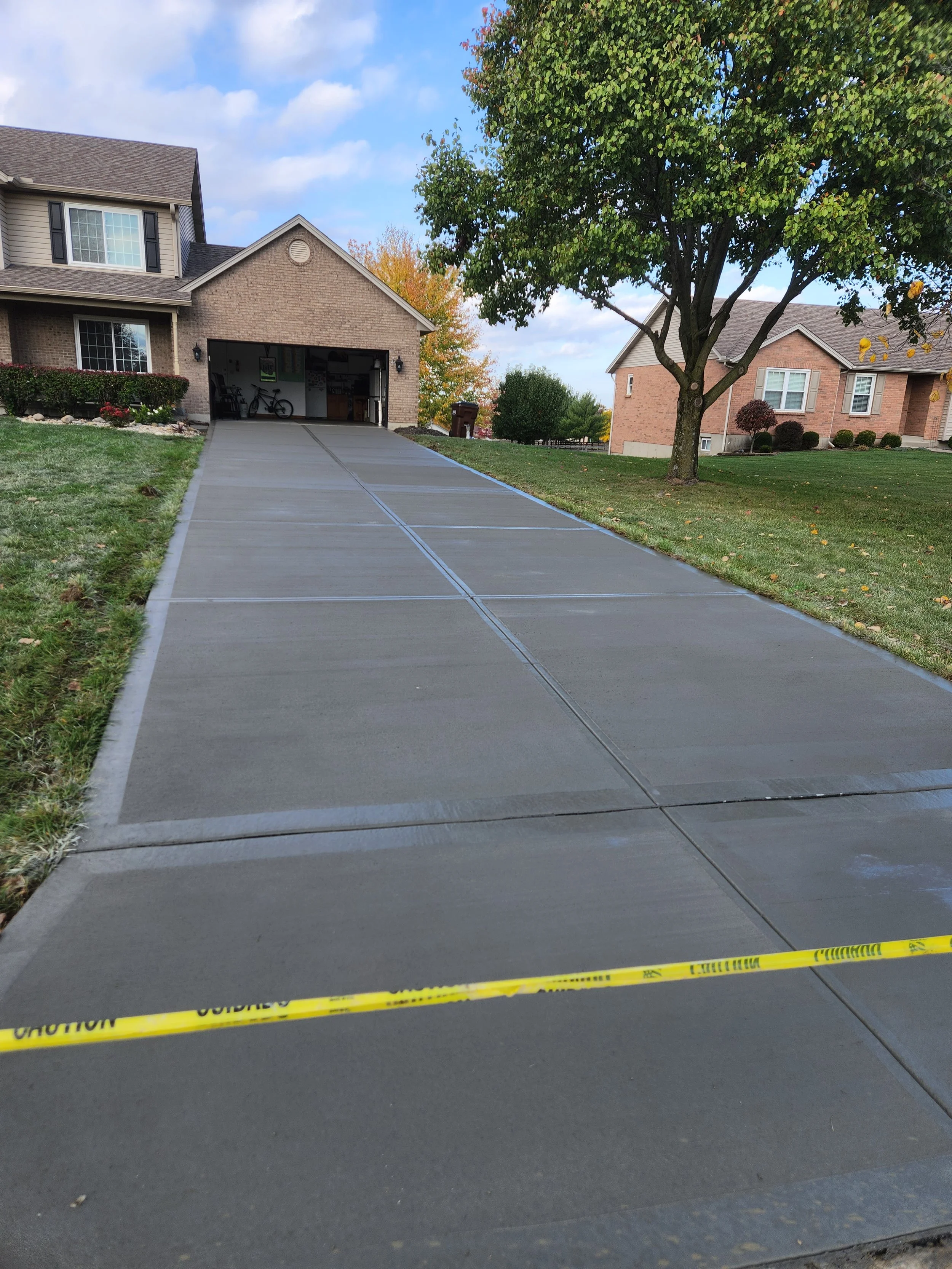 Freshly poured concrete driveway leading to a garage with a bike inside, surrounded by green lawns and trees in a suburban neighborhood.